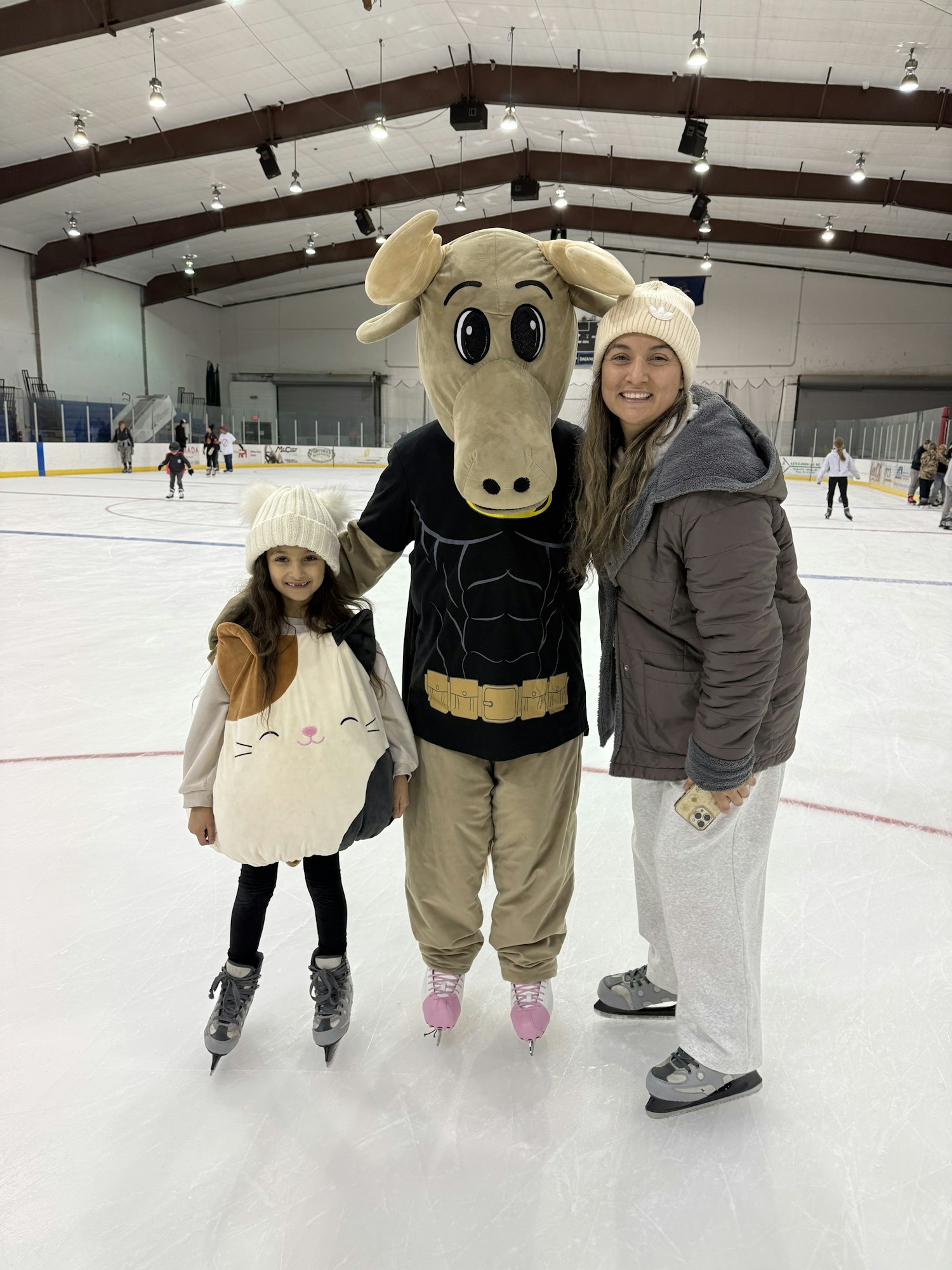 Two people and a mascot on ice skates, smiling in an ice rink setting, with a fun, cozy winter vibe.