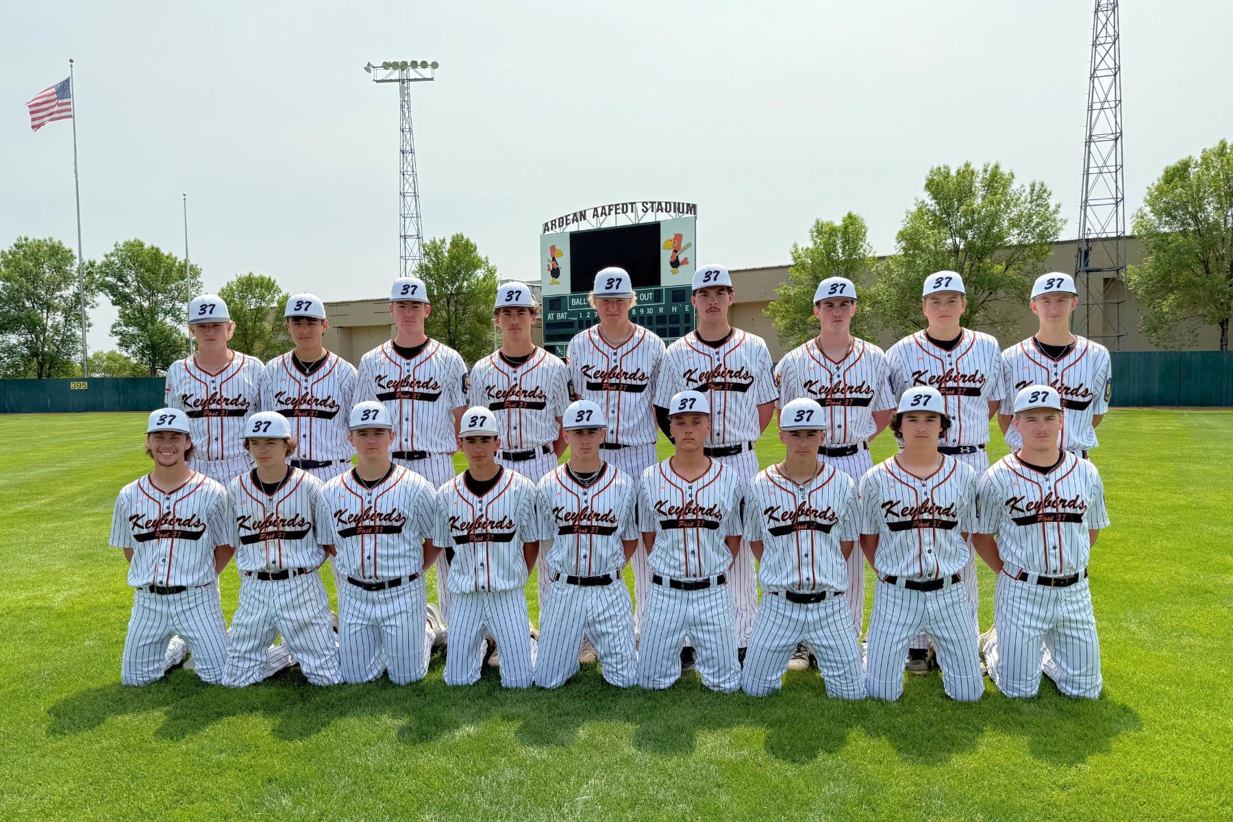 A baseball team poses in uniforms and hats with "37", on a field in front of a scoreboard and stadium backdrop.