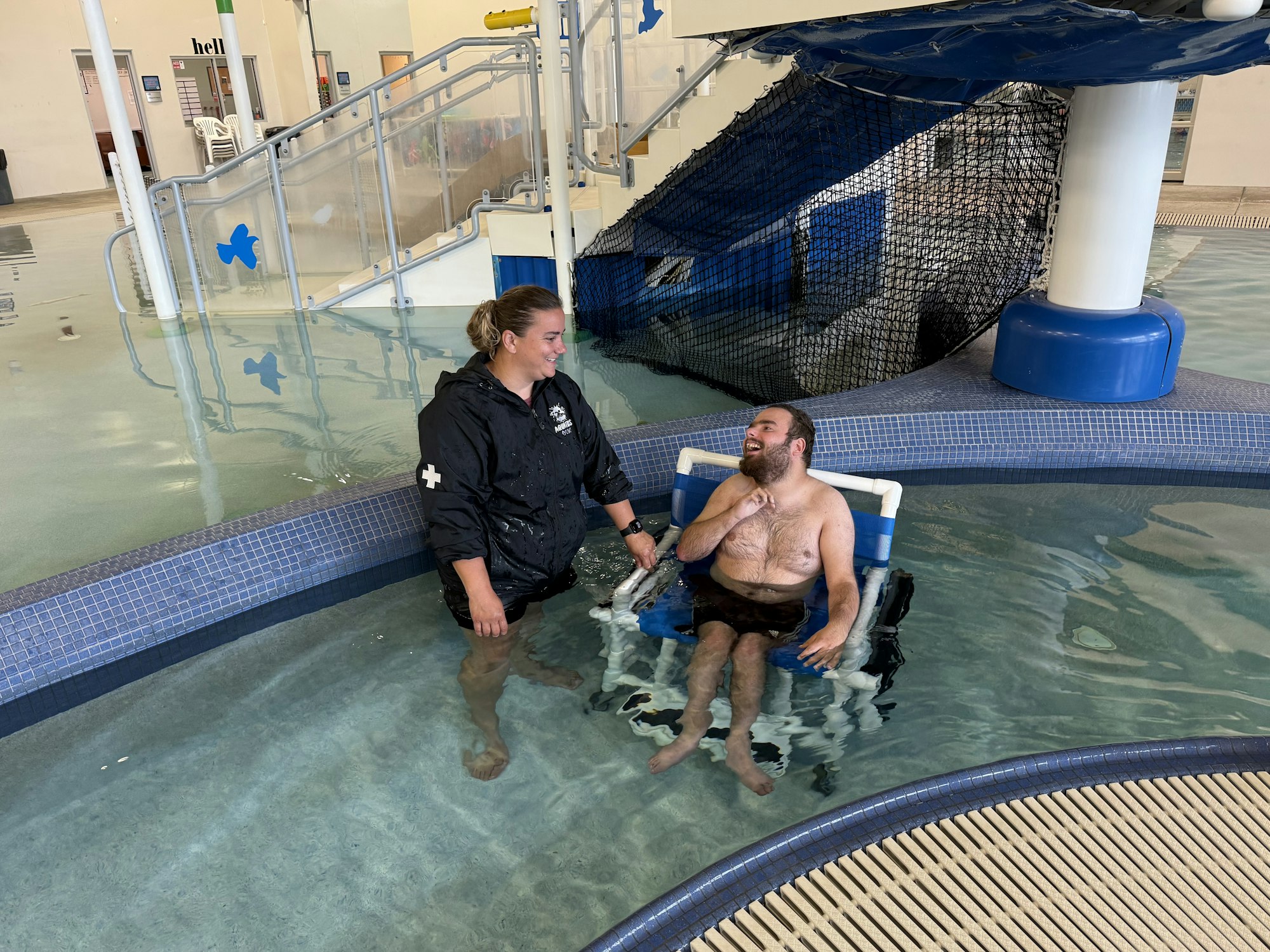 A person in a wheelchair in a pool is talking to a standing lifeguard.