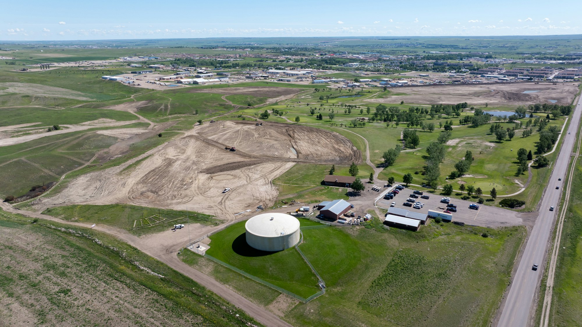 Aerial view of a landscape with construction activity, green fields, a water tank, and nearby buildings and roads.