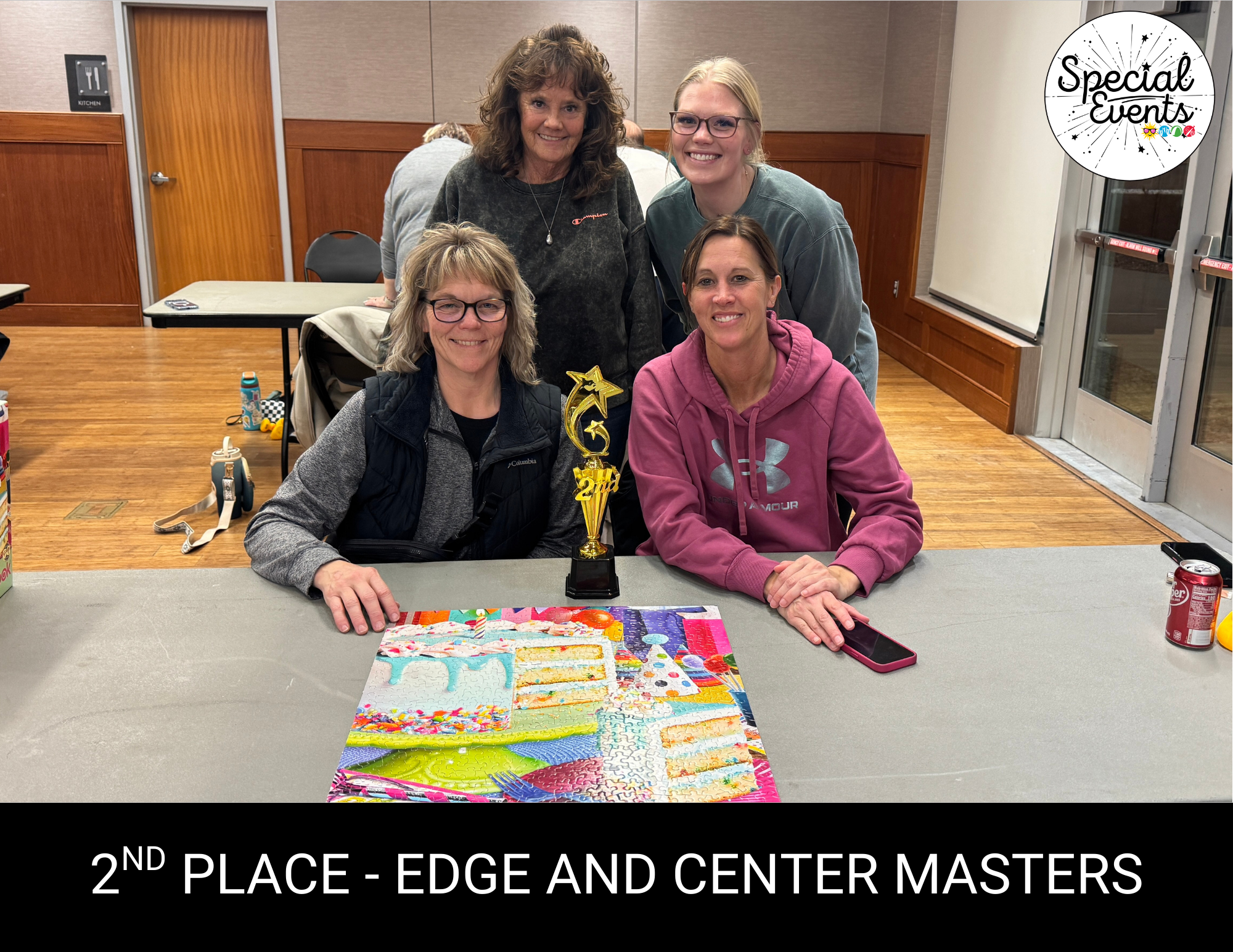 Four women proudly pose with a trophy for 2nd place at an event, beside a colorful puzzle on a table.