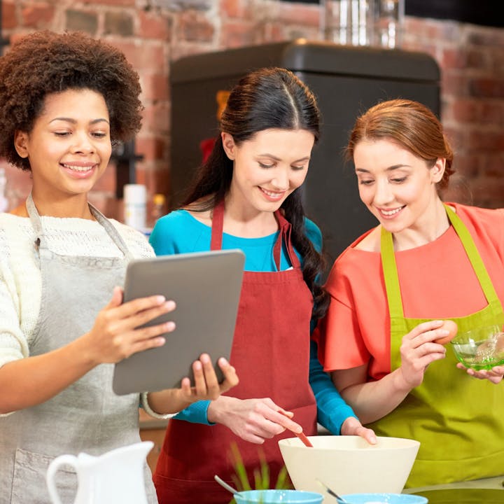Three women in aprons are cooking together in a kitchen, looking at a tablet while preparing food.