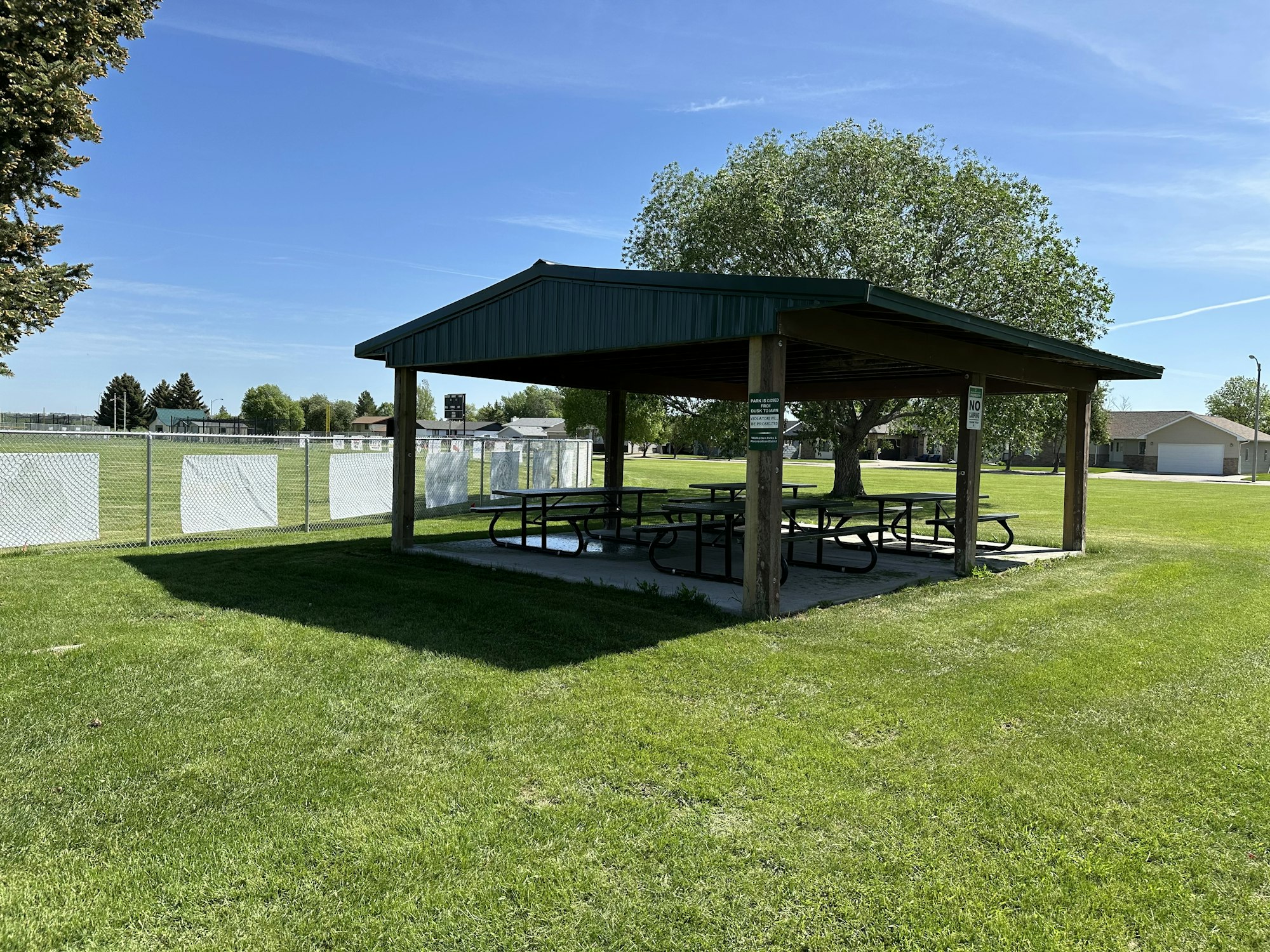 Park shelter with picnic tables, green grass, trees, and a chain-link fence in a sunny setting.