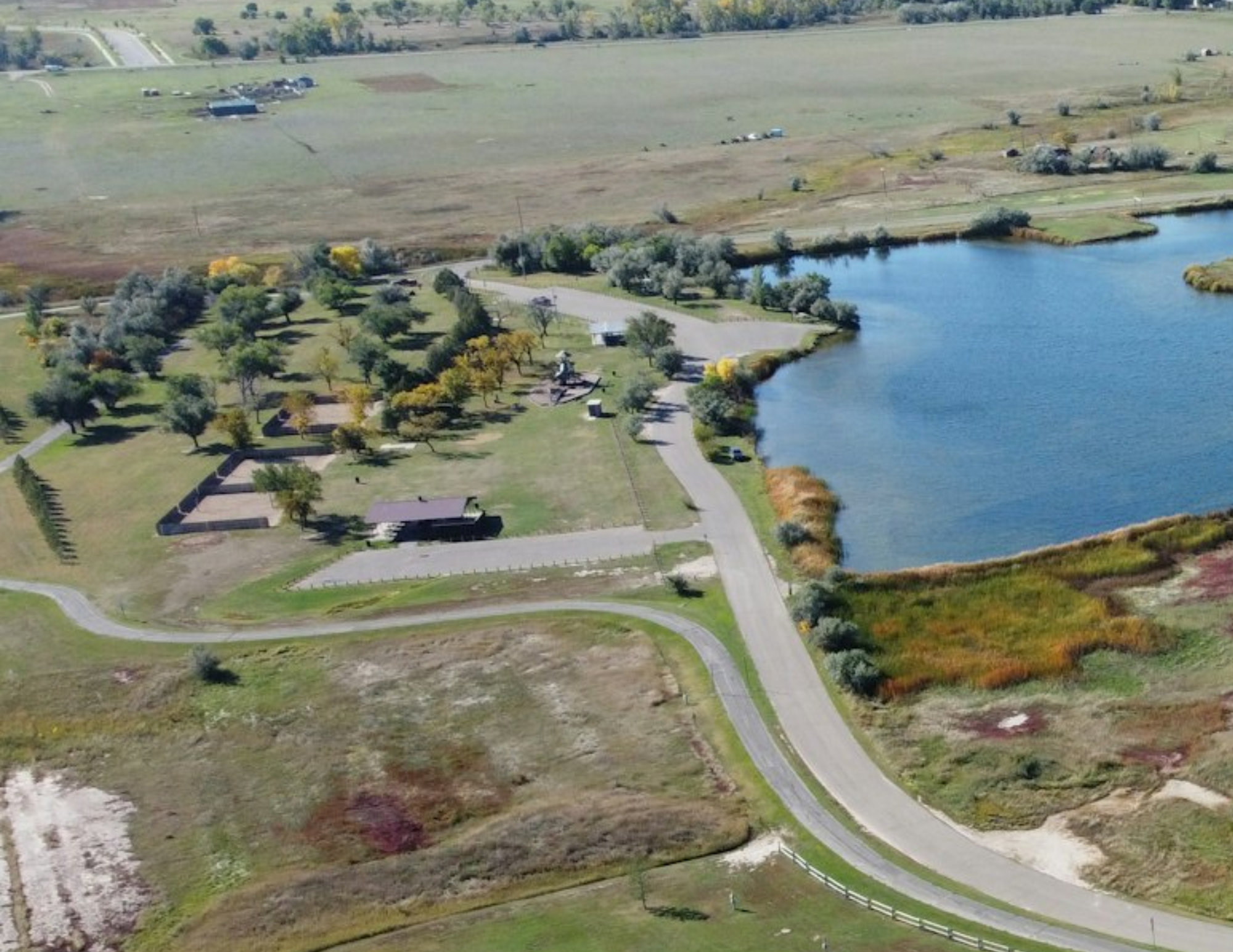 Aerial view of a park with a lake, trees, paths, a building, and open fields.