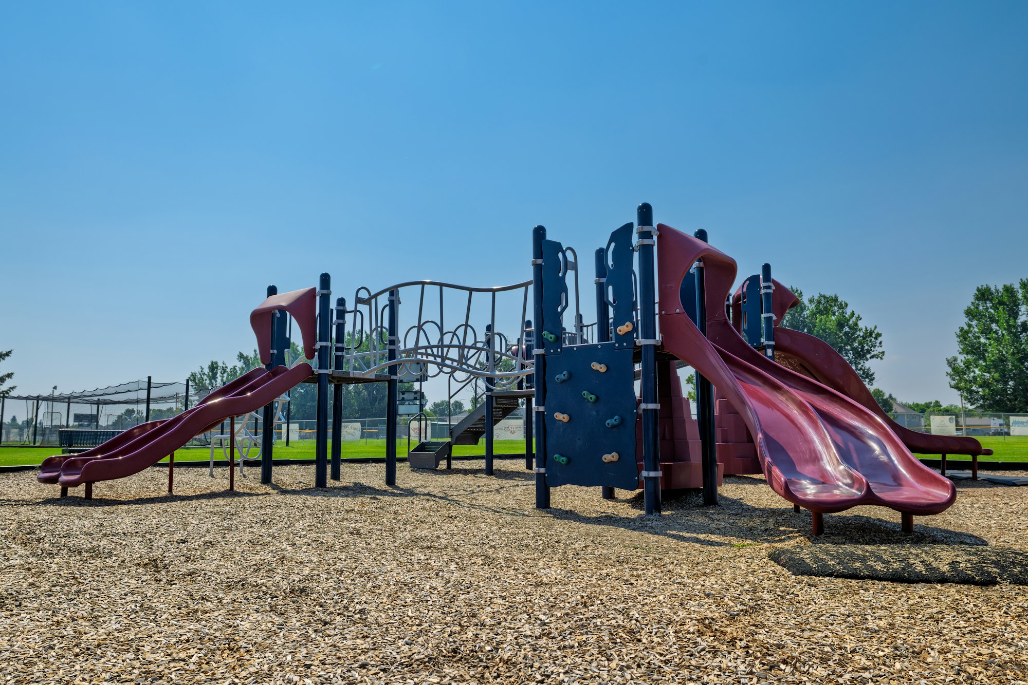 A playground structure with two slides, climbing wall, and safety surfacing, set against a clear blue sky and green grass.