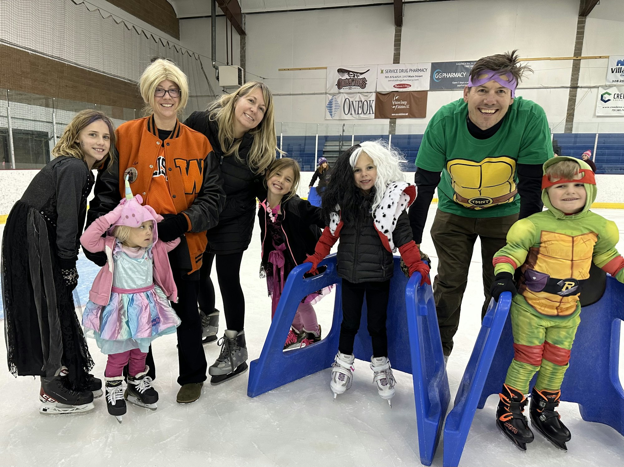 A group of people with some in costumes ice-skating, using skate aids for support. There's a happy and festive atmosphere.