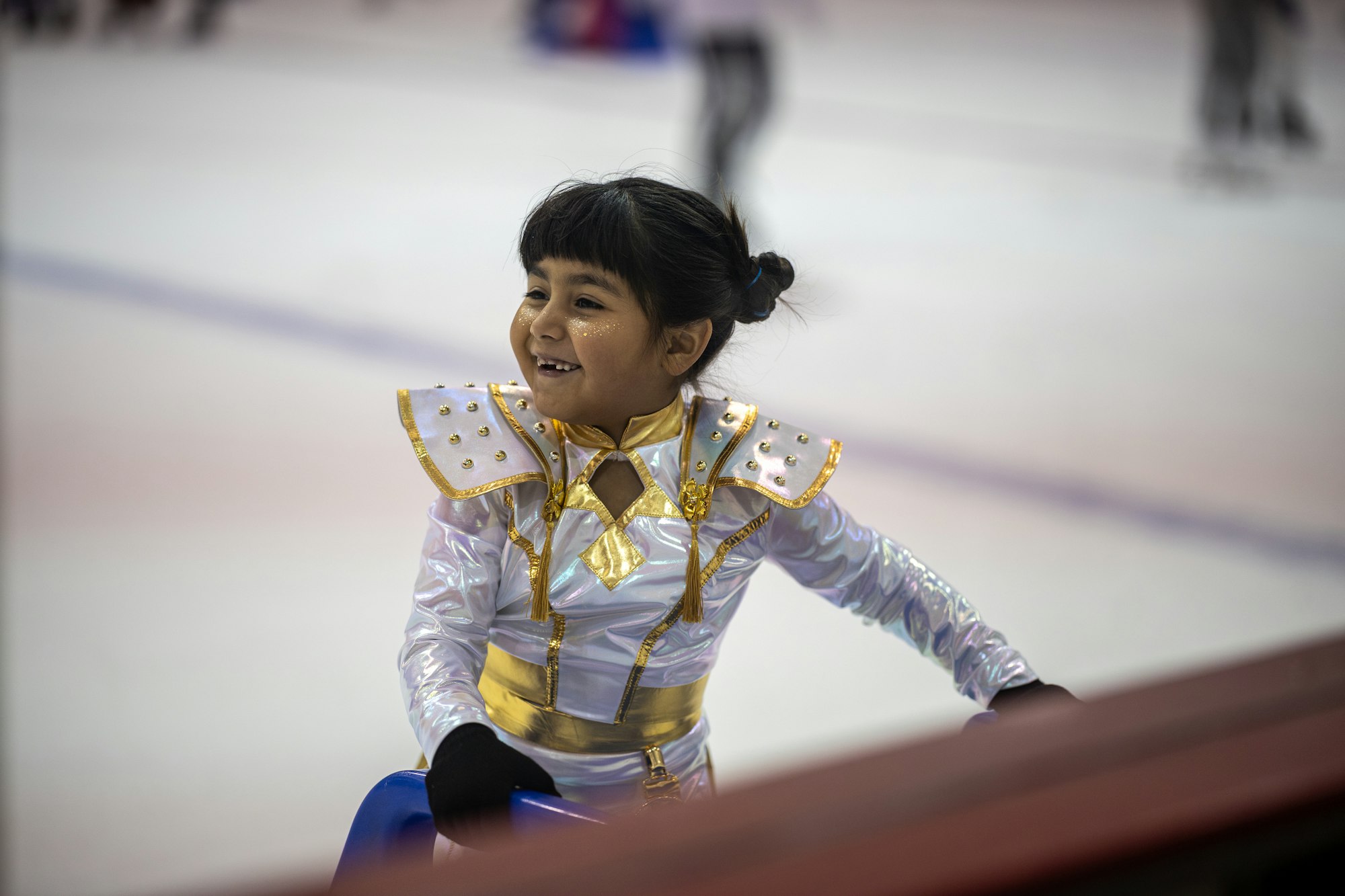 A young child in a shiny, gold-accented skating outfit smiles while holding onto a blue skating aid on the ice rink.