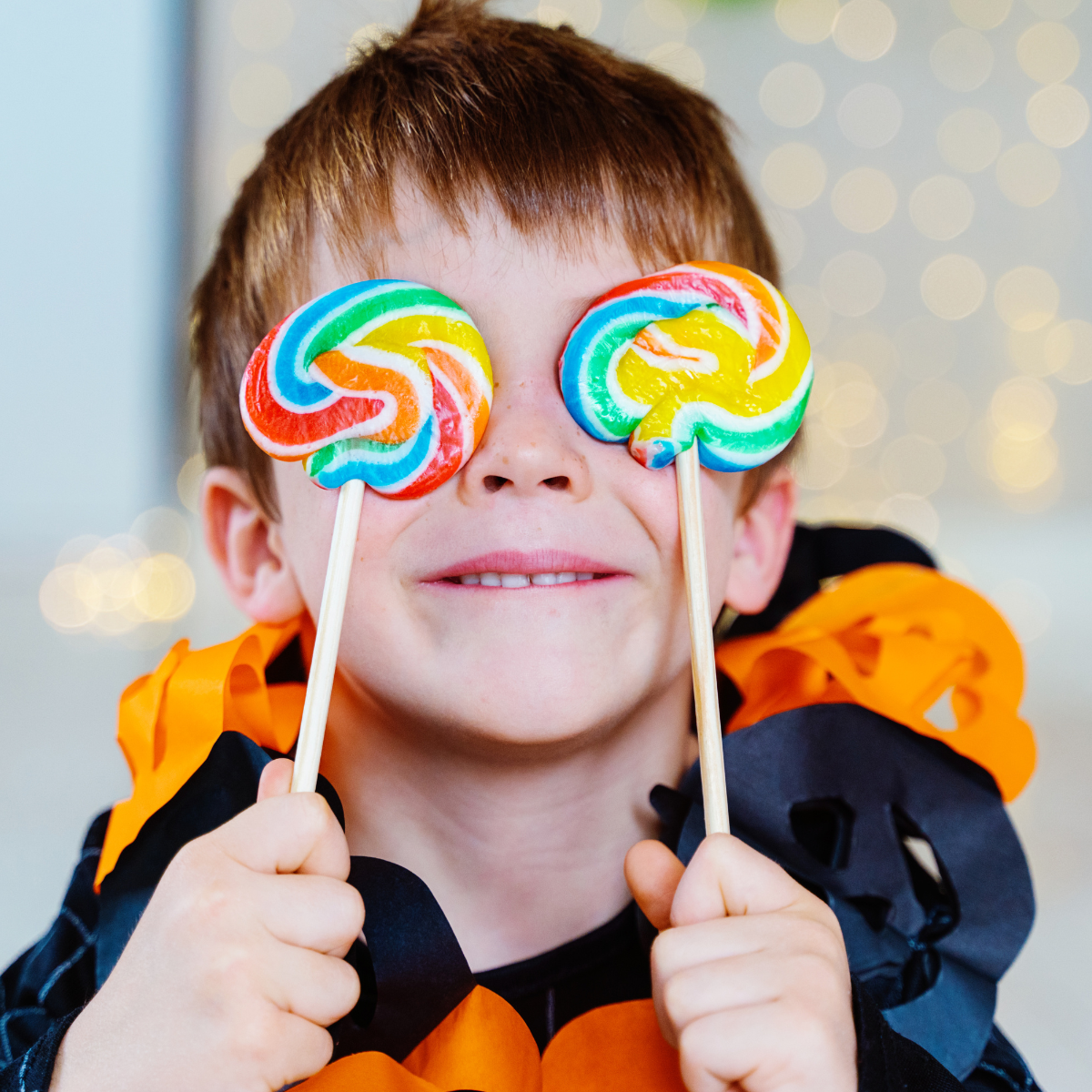 A happy child holds colorful lollipops over his eyes, wearing a festive black and orange costume, with bright lights in the background.