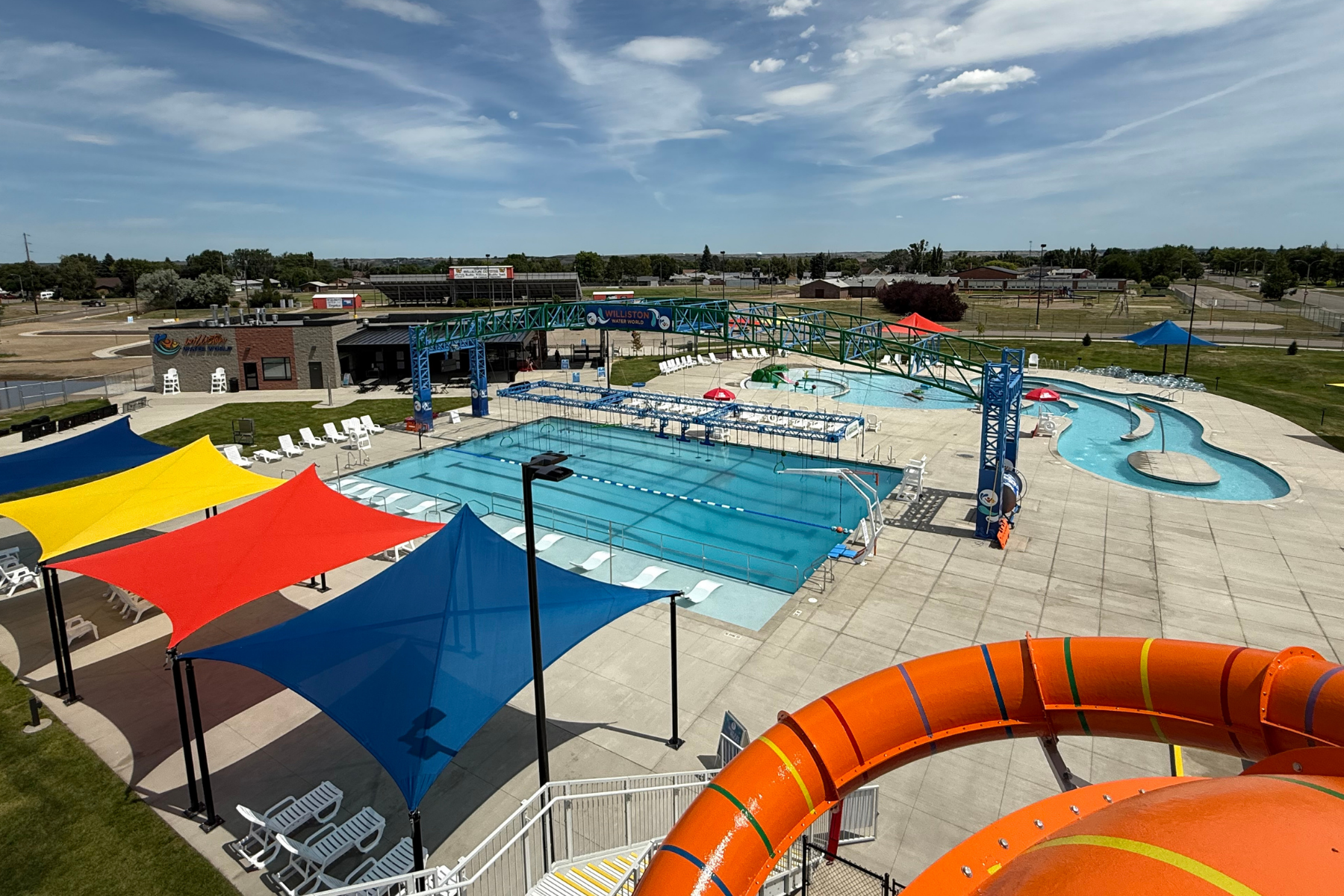 The image shows a colorful outdoor swimming pool area with water slides, shaded areas, and landscaped surroundings under a blue sky.ld