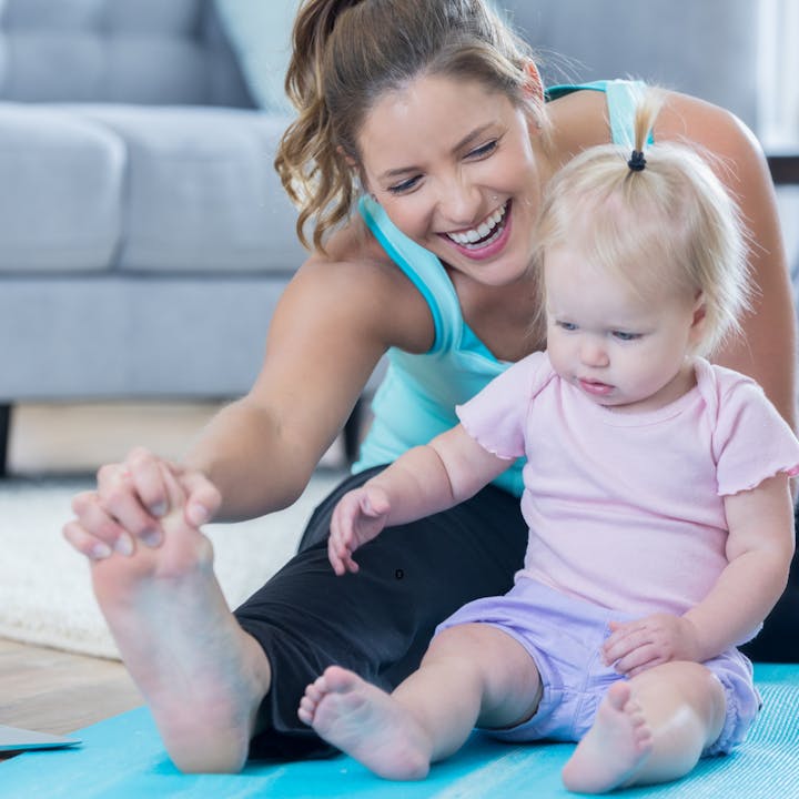 A woman and a baby sitting on a yoga mat, smiling and stretching together.