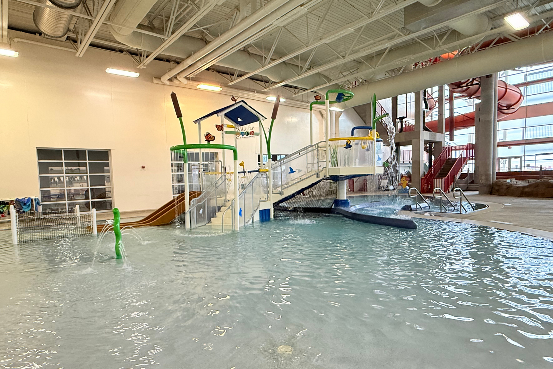 An indoor water play area features a slide, splash pads, and pools with playful decorations and a water slide in the background.