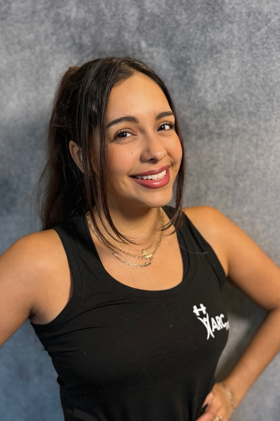 A person smiling, wearing a black sleeveless top with a logo, against a gray background.