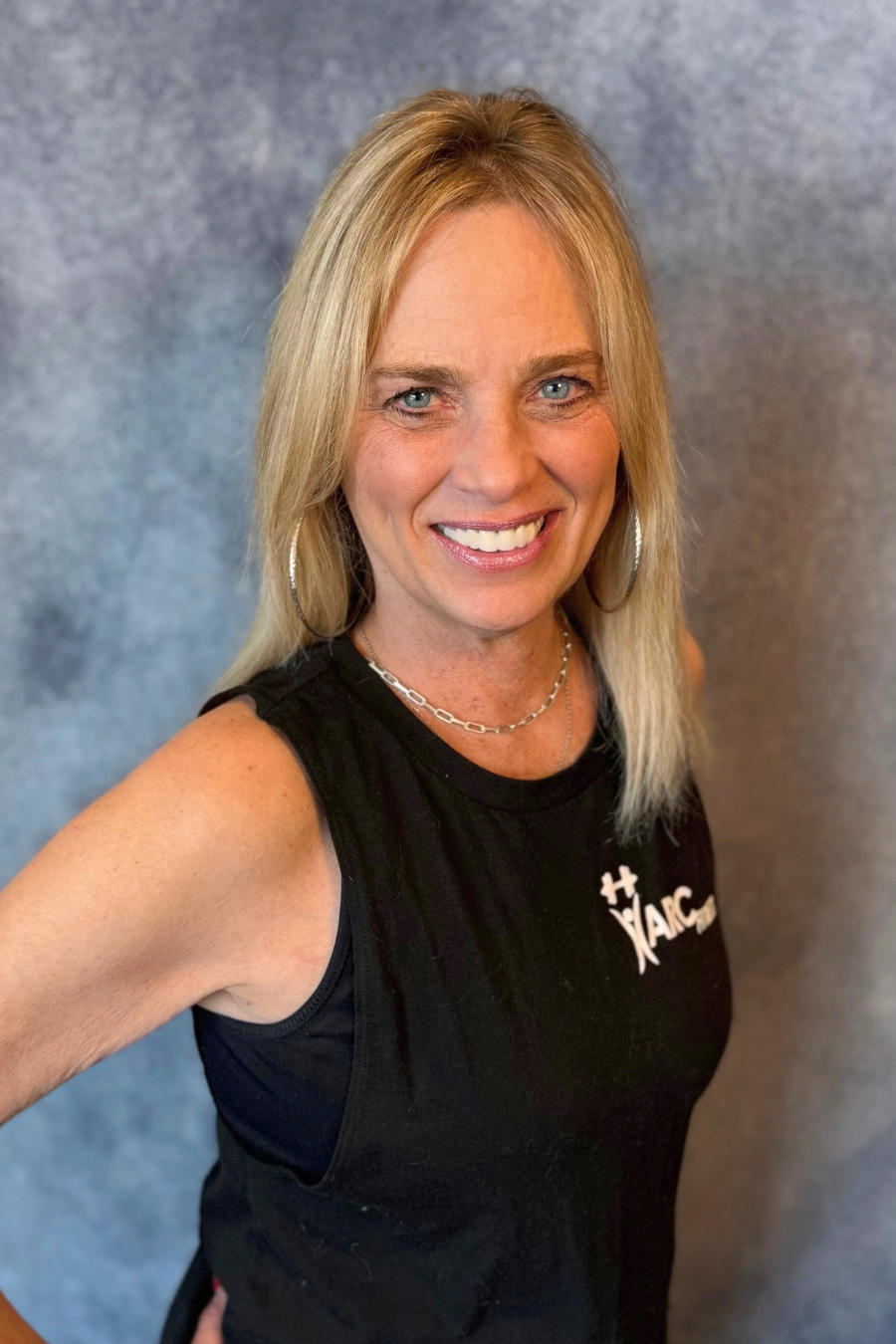A woman with blonde hair smiling, wearing a black sleeveless top with a logo, in front of a gray backdrop.