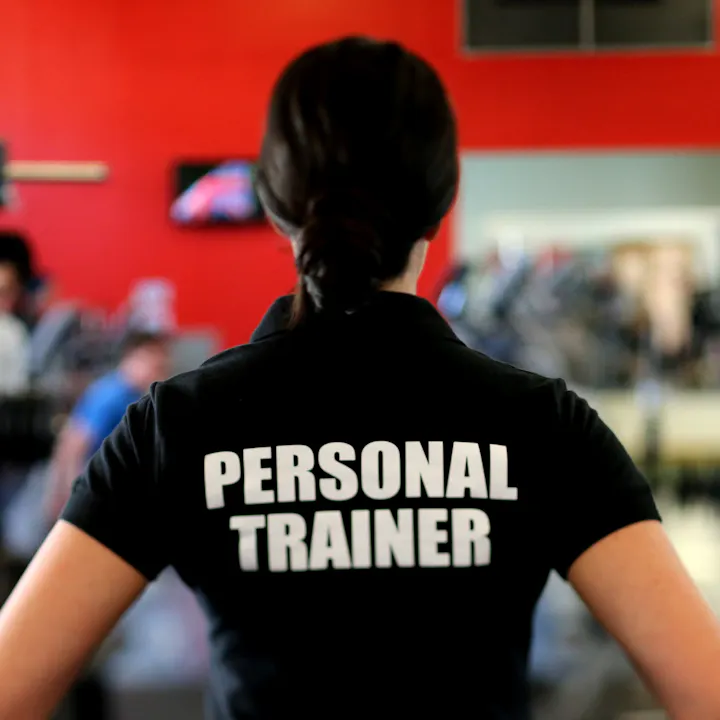 A person wearing a black shirt with "PERSONAL TRAINER" printed on the back, standing in a gym environment.