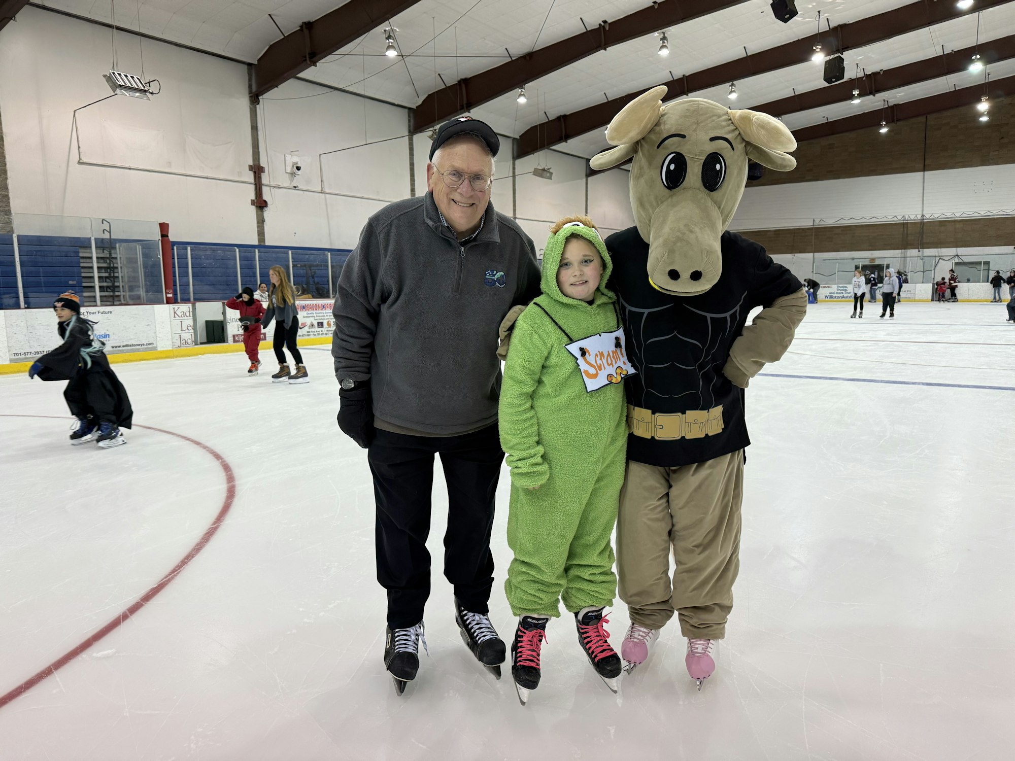 A group of skaters at an ice rink, featuring a man, a child in a green costume, and a person in a moose mascot outfit.