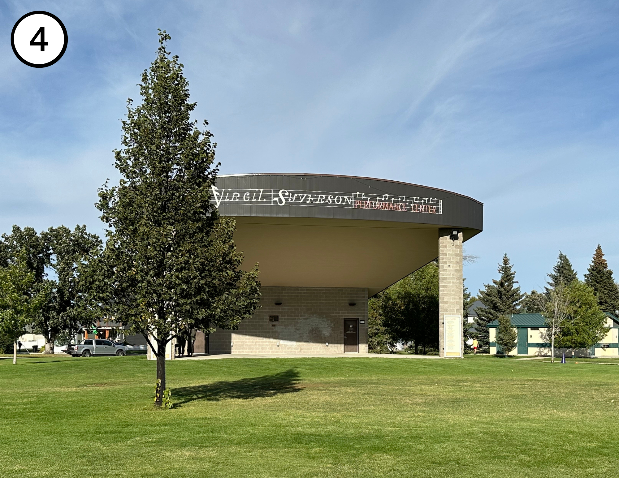 Outdoor performance center with a curved roof, grassy area, and trees; one car in the background.