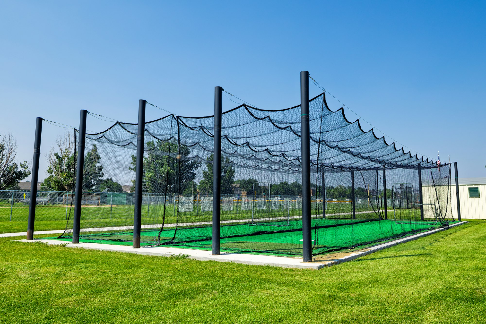 The image shows a batting cage with black nets set up on a green field under a clear blue sky.