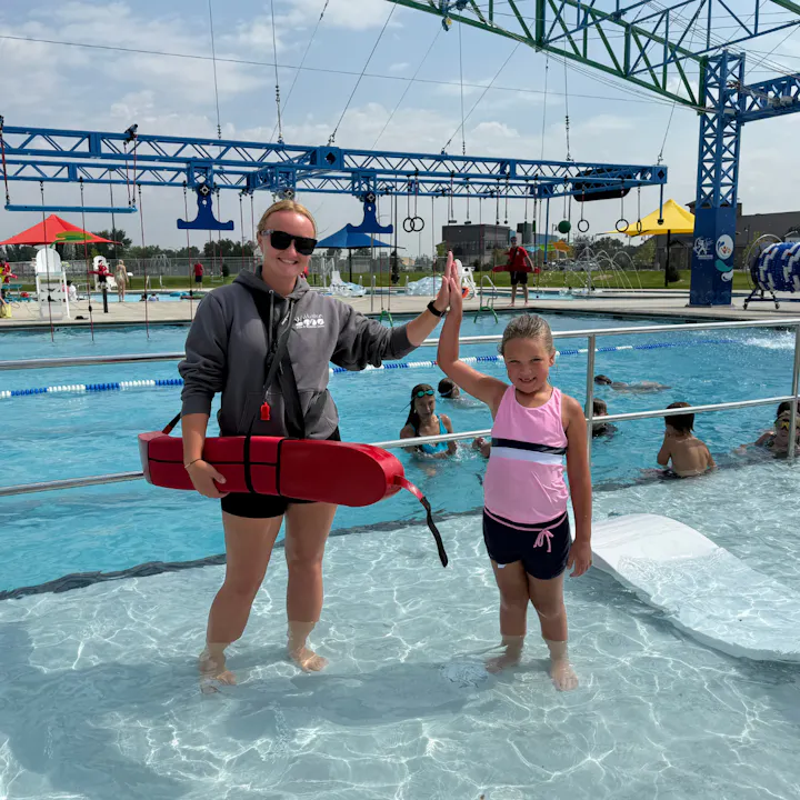 A lifeguard and a young girl high-five in a swimming pool, surrounded by other children enjoying the water activities.