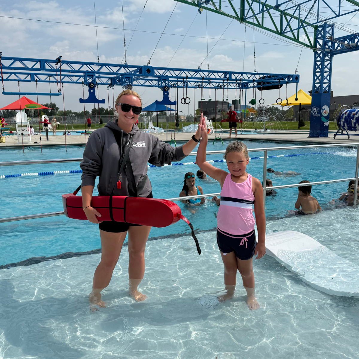 A lifeguard and a young girl high-five in a swimming pool, surrounded by other children enjoying the water activities.