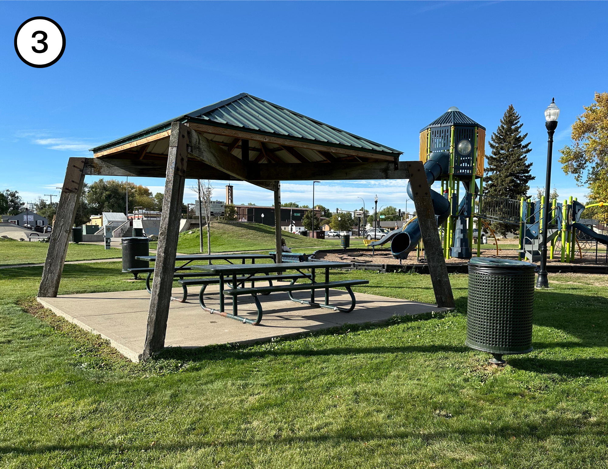 A park with a picnic shelter, a playground structure, a trash can, and a lamppost on a sunny day.