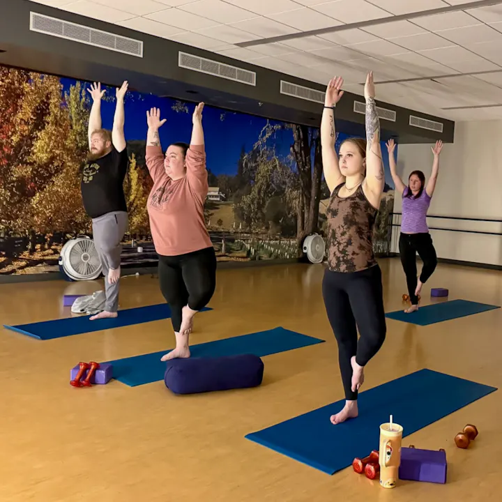 Four people practicing yoga in a studio, each balancing on one leg with arms raised. Yoga mats and props are visible.