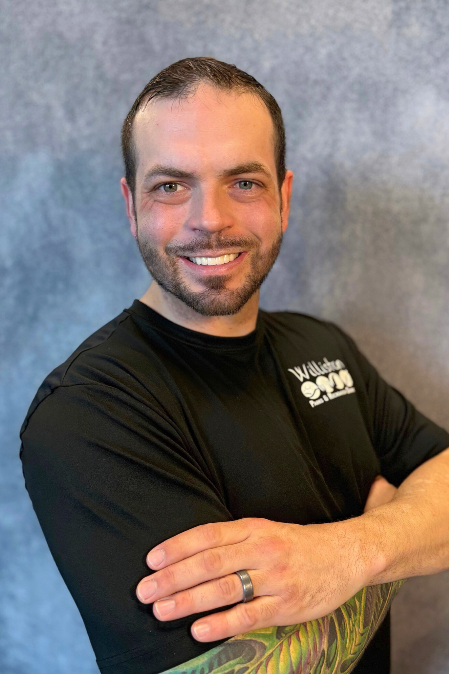 A person smiling, arms crossed, wearing a black shirt and showing a colorful arm tattoo.