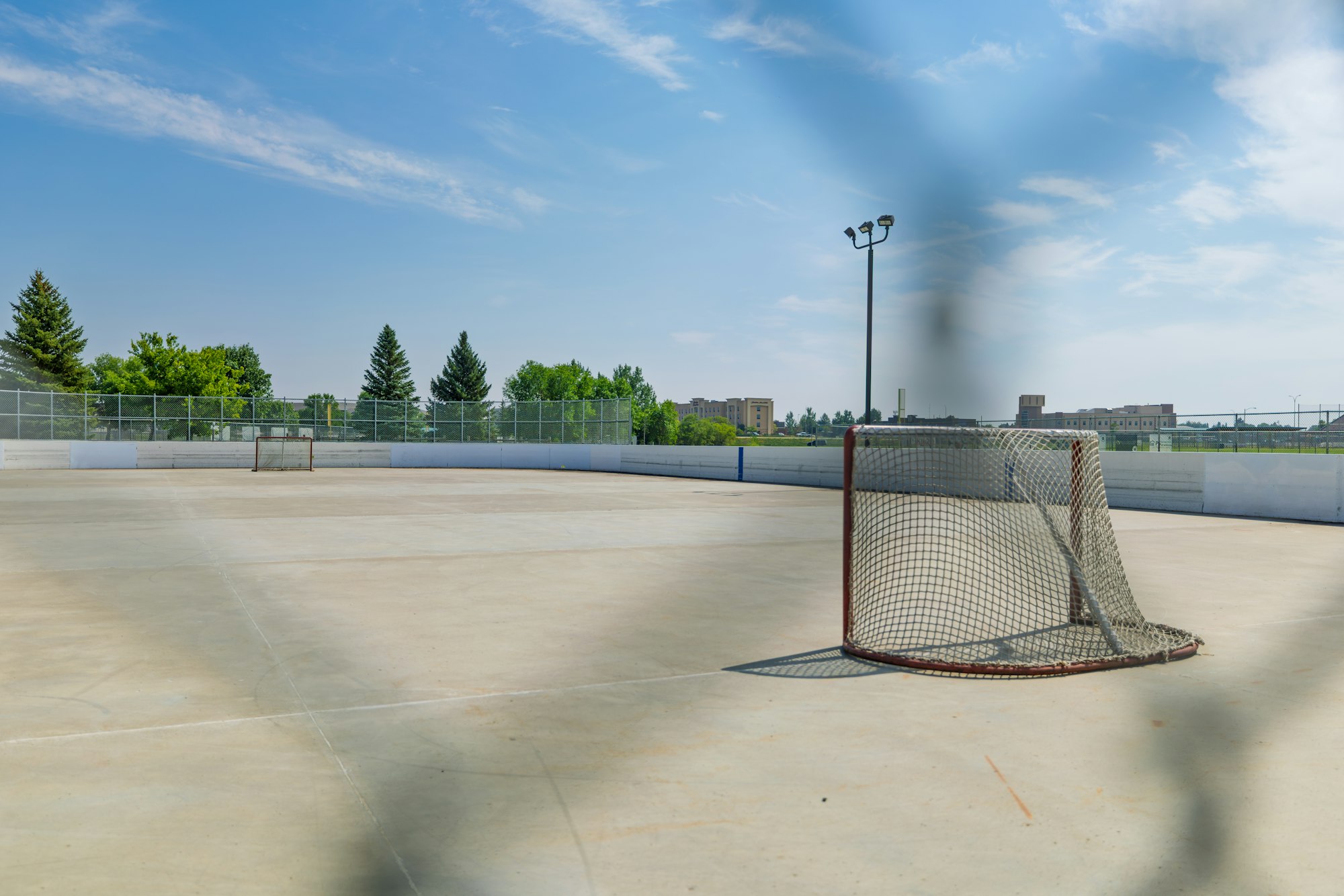 An empty outdoor hockey rink with two goal nets, surrounded by trees and a blue sky.