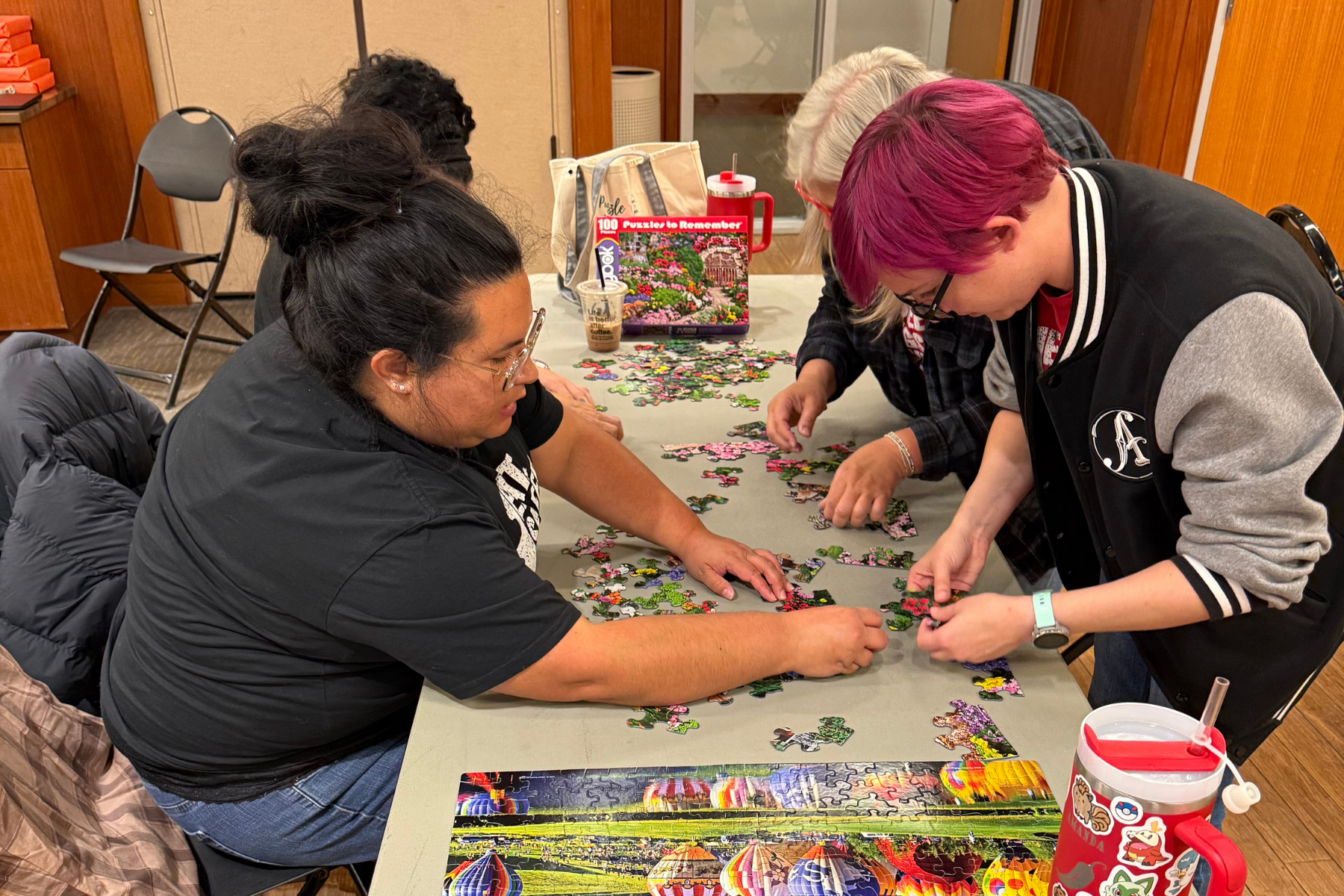 A group of people is assembling a colorful puzzle at a table, with pieces scattered around and some drinks nearby.