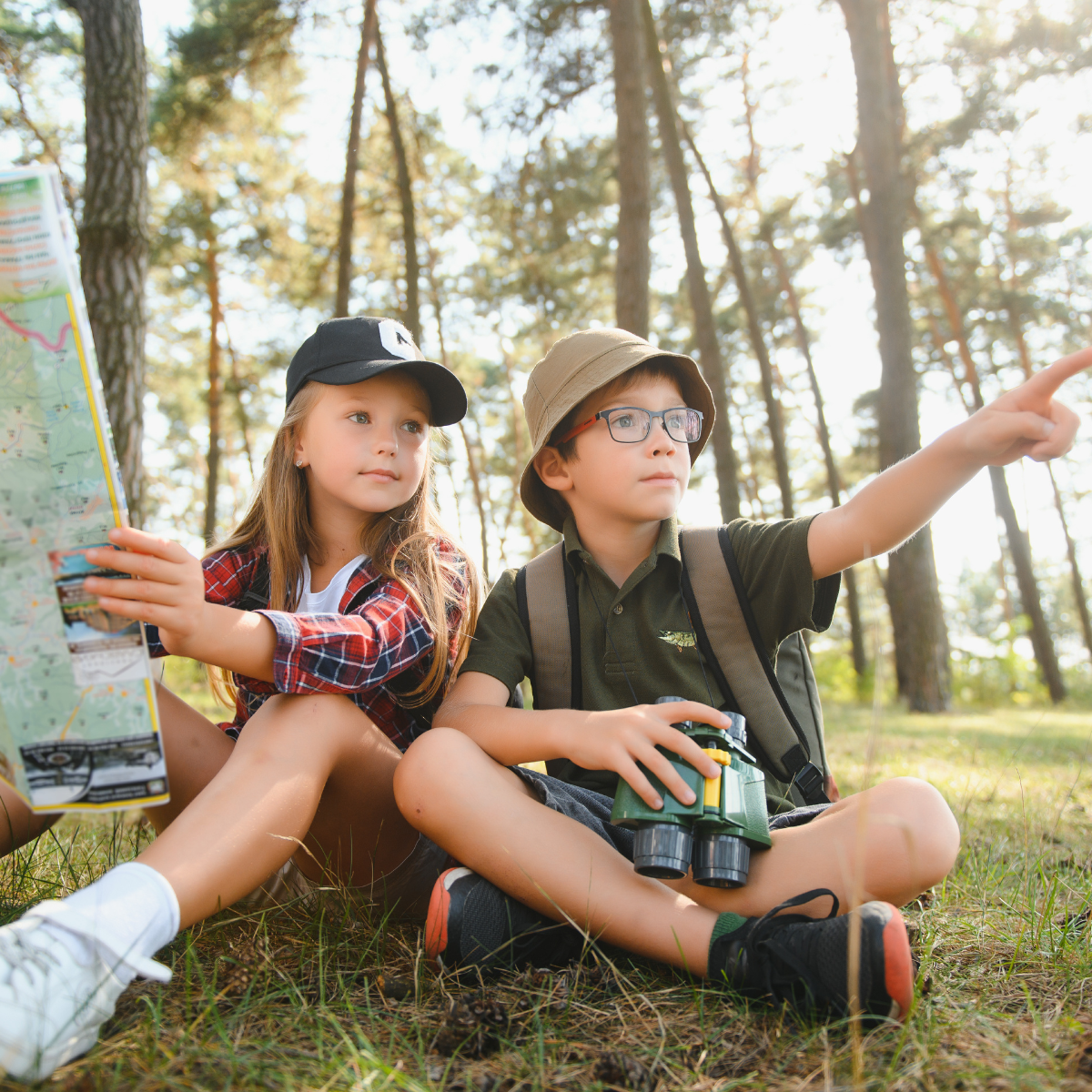Two kids are sitting in a forest, studying a map and exploring with binoculars, enjoying a day of outdoor adventure.