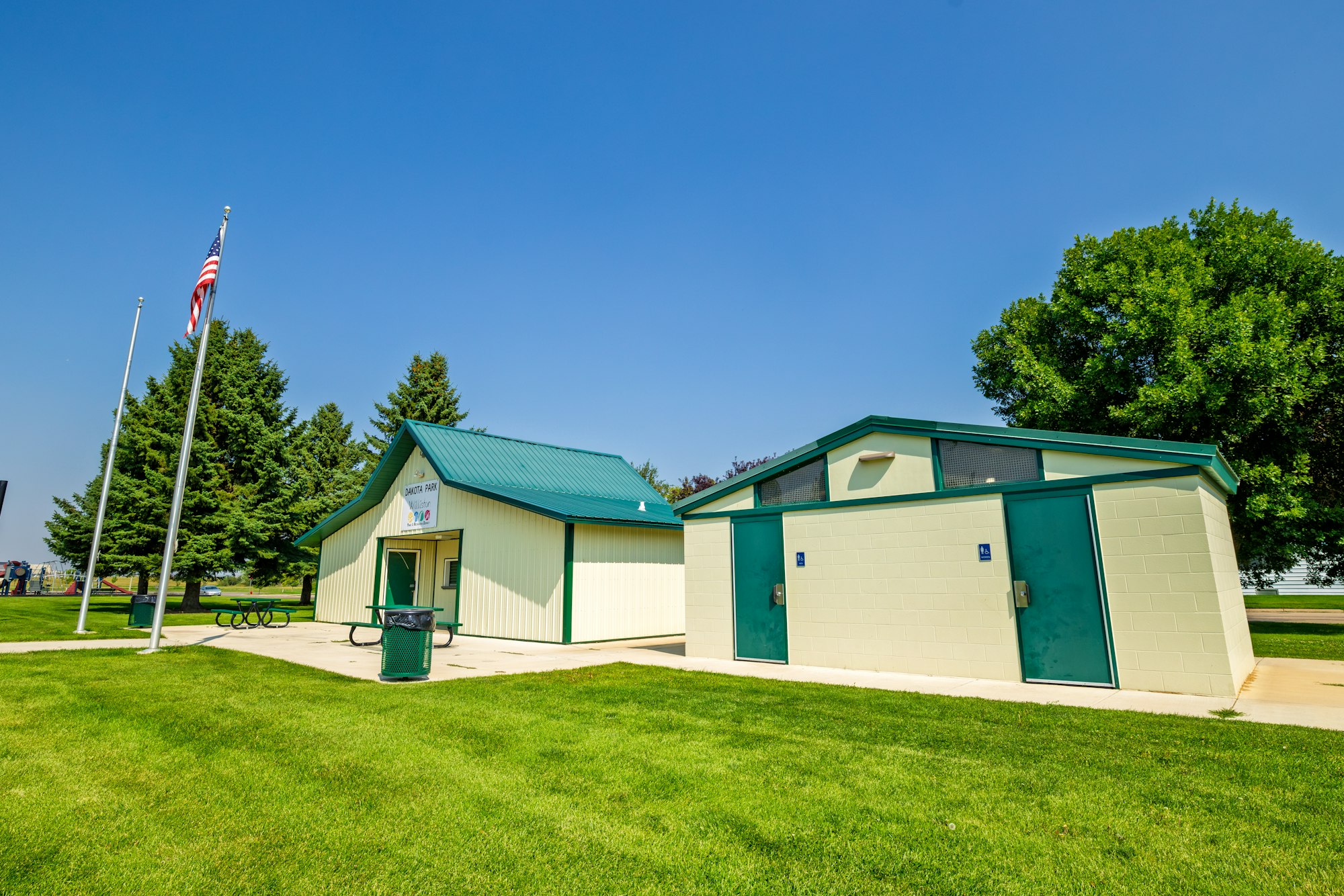 A park building with a green roof, restrooms, a grassy area, and American flags nearby on a clear blue sky day.