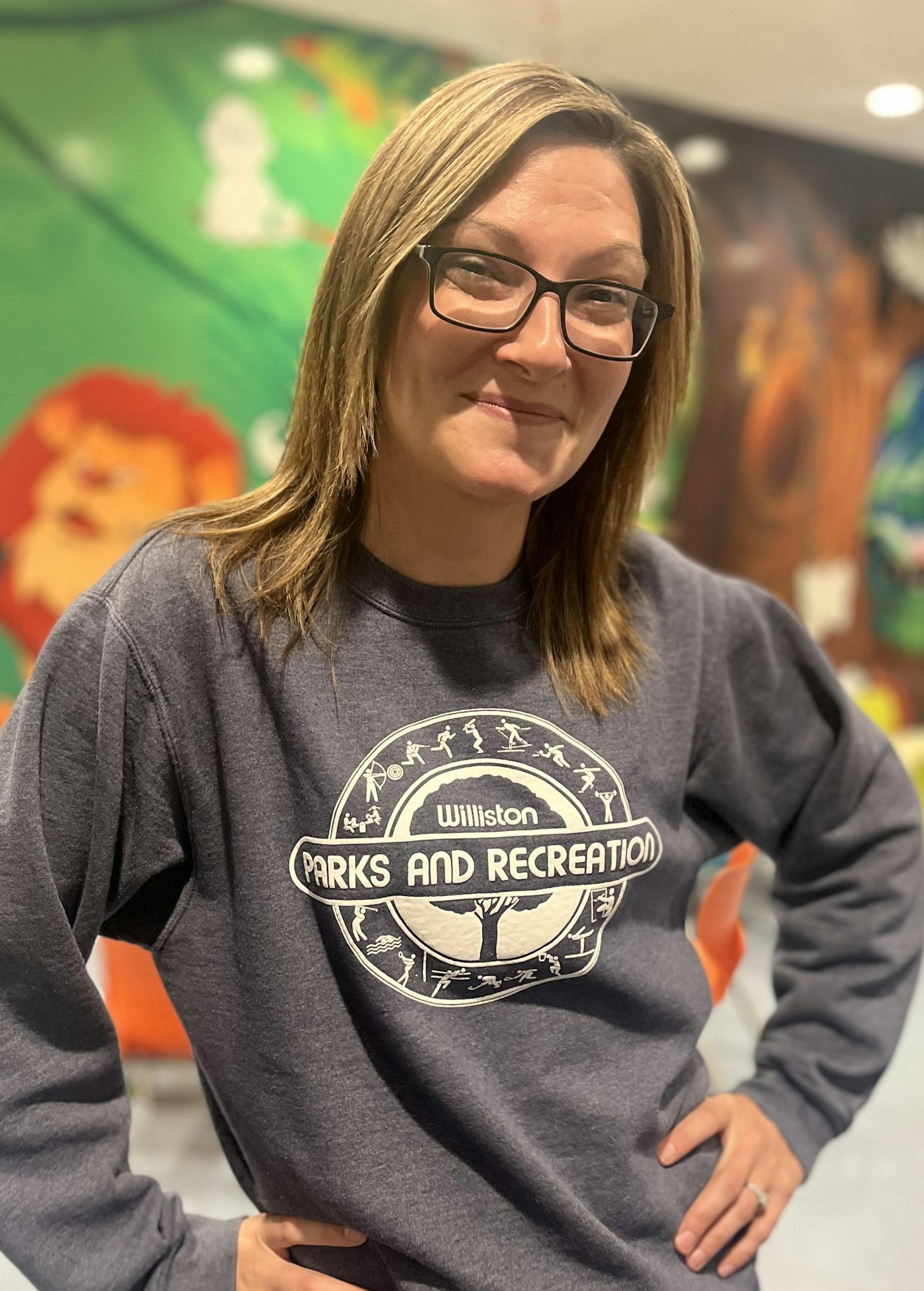 A smiling person wearing glasses and a "Parks and Recreation" sweatshirt, standing in front of a colorful wall with cartoon characters.