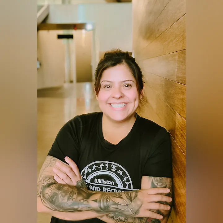 A smiling person with tattoos is posing with arms crossed against a wooden wall in a well-lit indoor space.