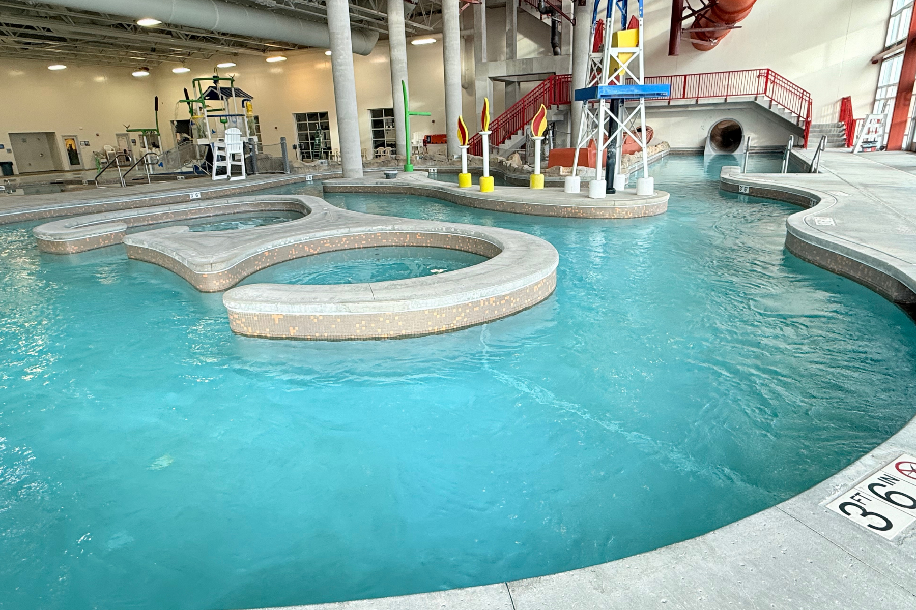 The image shows a modern indoor water park area with pools, water slides, and play structures in a brightly lit space.