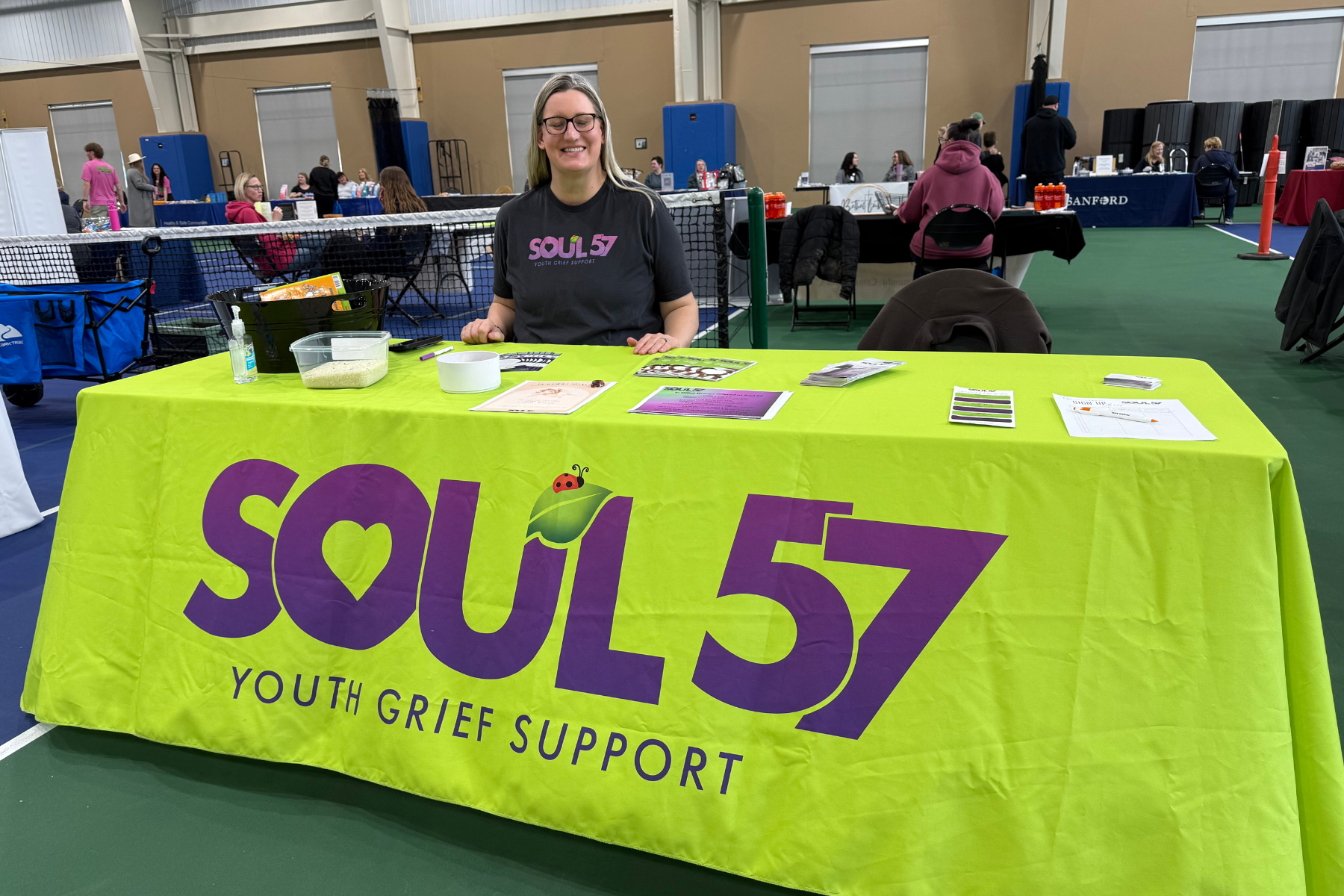 A smiling woman at a booth showcasing "Soul 57," a youth grief support organization, with informational materials displayed.