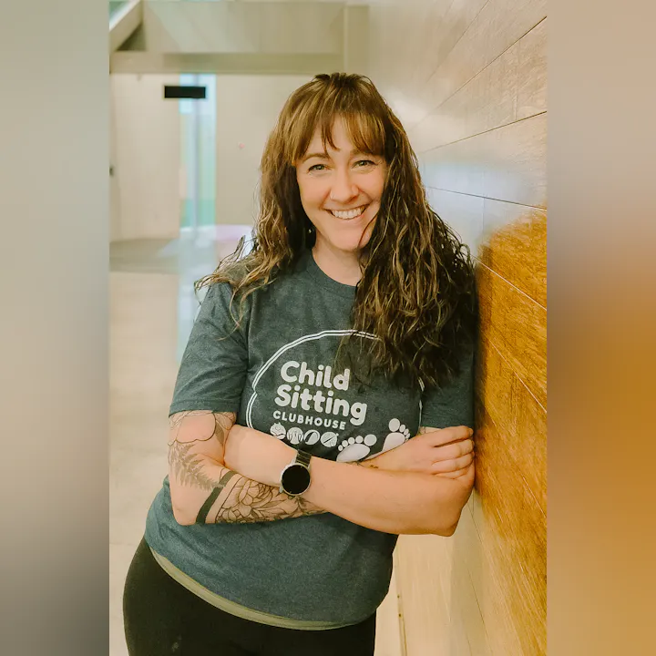 A smiling person with curly hair stands against a wooden wall, wearing a "Child Sitting Clubhouse" t-shirt.