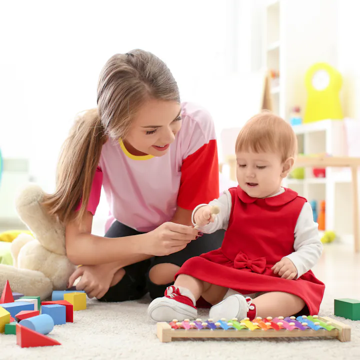 A woman and a baby play with colorful toys on a rug in a bright room.