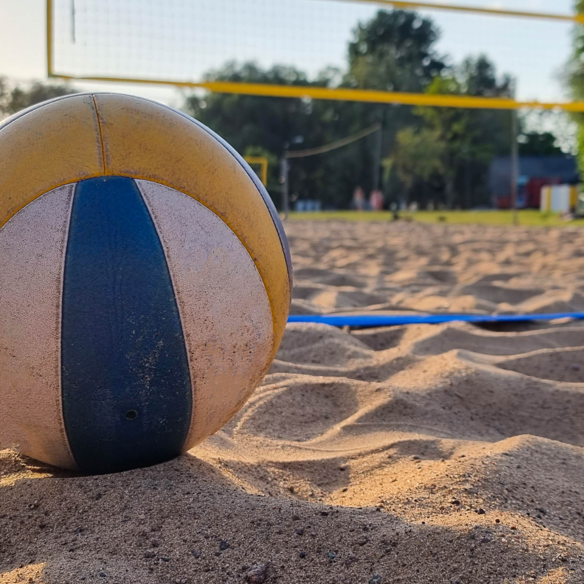 A close-up of a volleyball resting on sandy ground, with a volleyball net and trees in the background, hinting at a beach court.