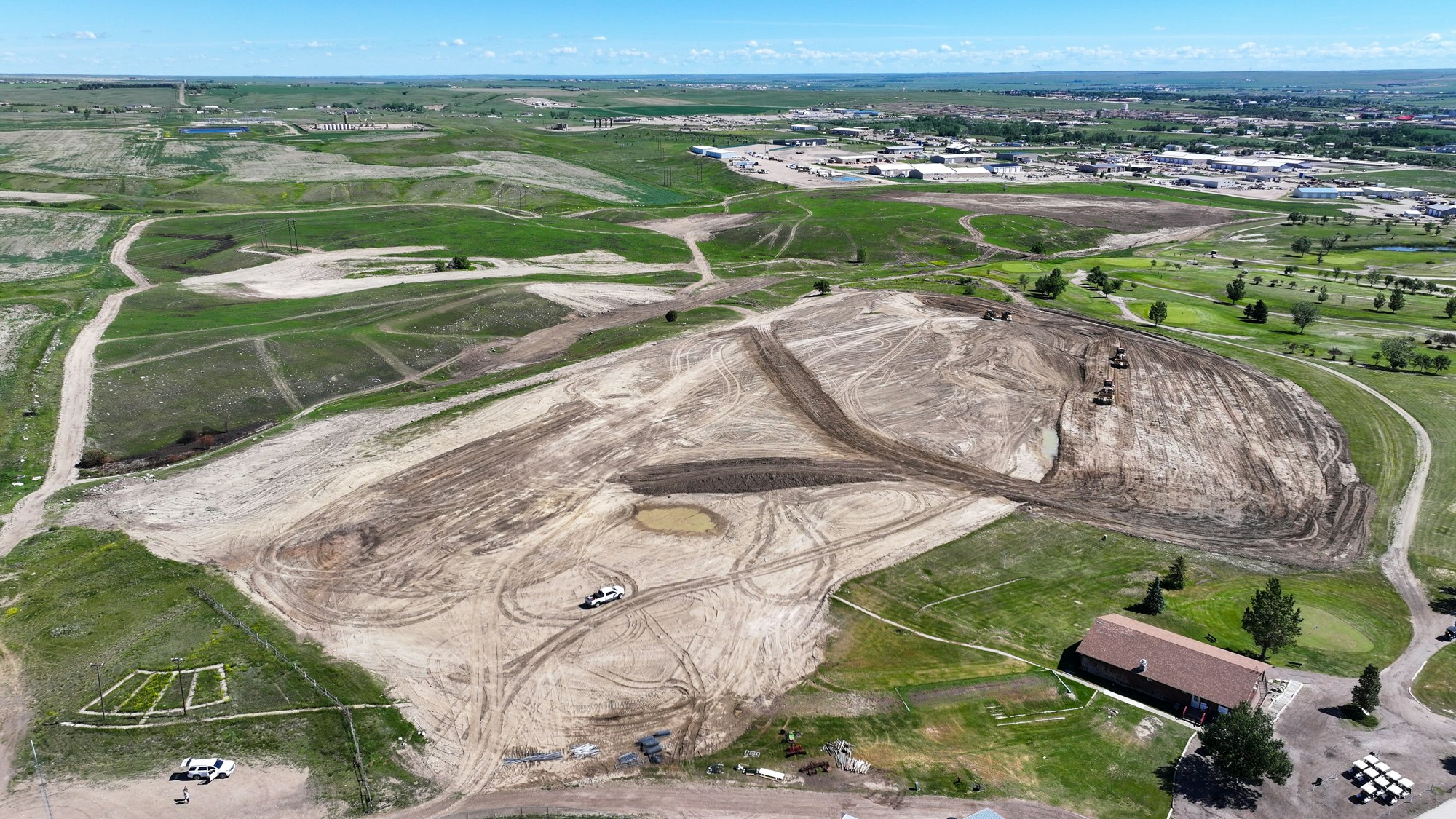 An aerial view of a construction site with cleared land, machinery working, and nearby developments in a green landscape.