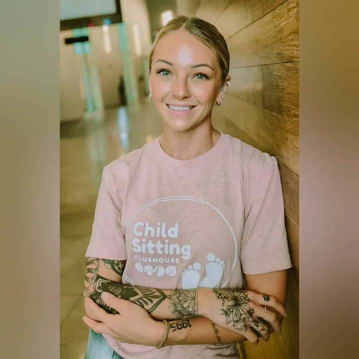 A smiling person with tattoos is sitting against a wooden wall, wearing a pink "Child Sitting Clubhouse" t-shirt.