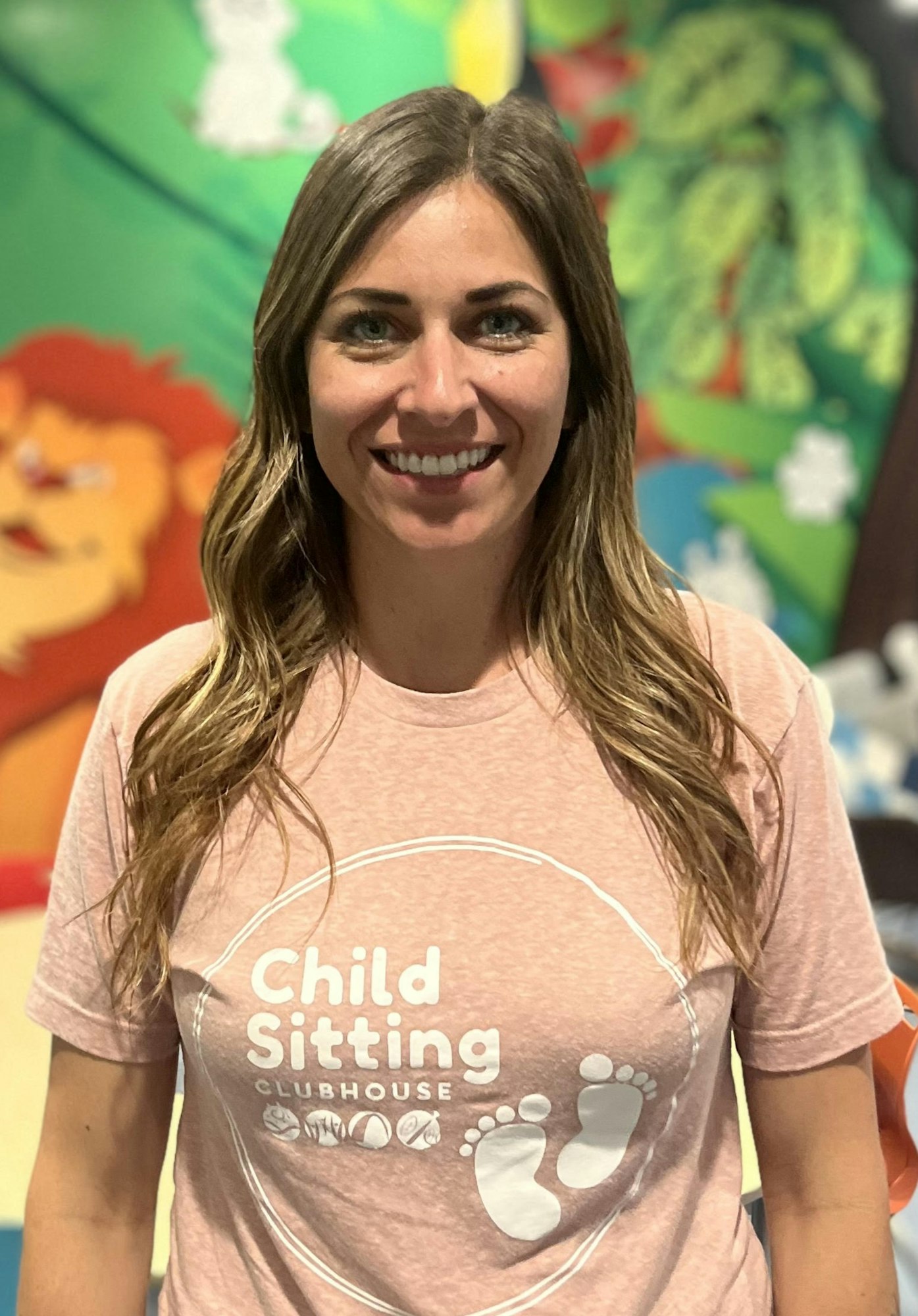 A smiling woman in a “Child Sitting Clubhouse” t-shirt, with colorful murals in the background.