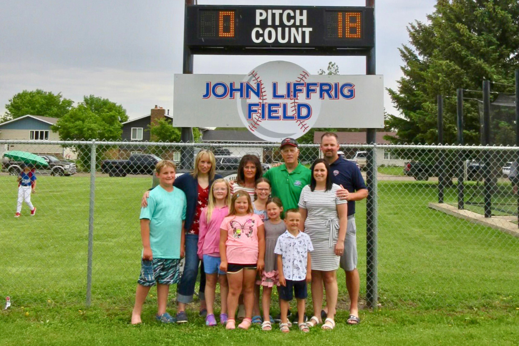 A group of people posing for a photo at "John Liffreig Field" with a baseball scoreboard in the background.