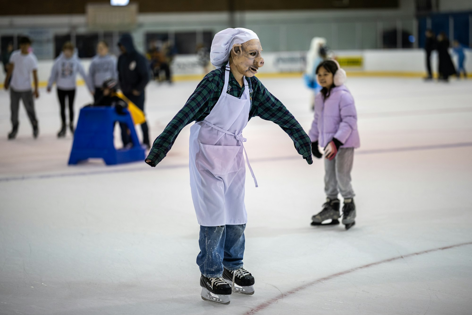An individual wearing a pig mask and apron is ice skating, surrounded by other skaters in an ice rink.