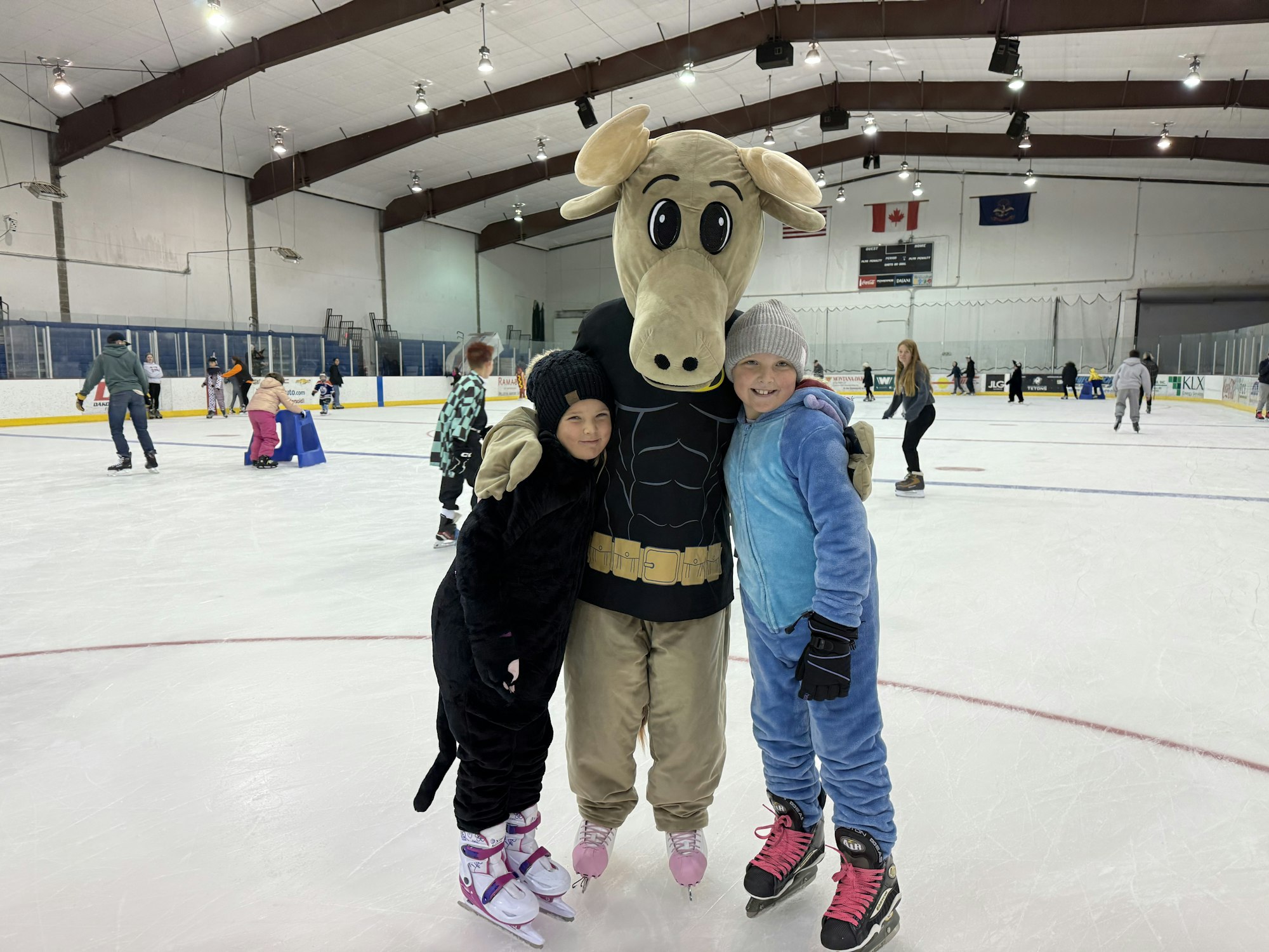 Children ice skating with a person in a moose costume at a rink, with others skating in the background. Fun winter activity!