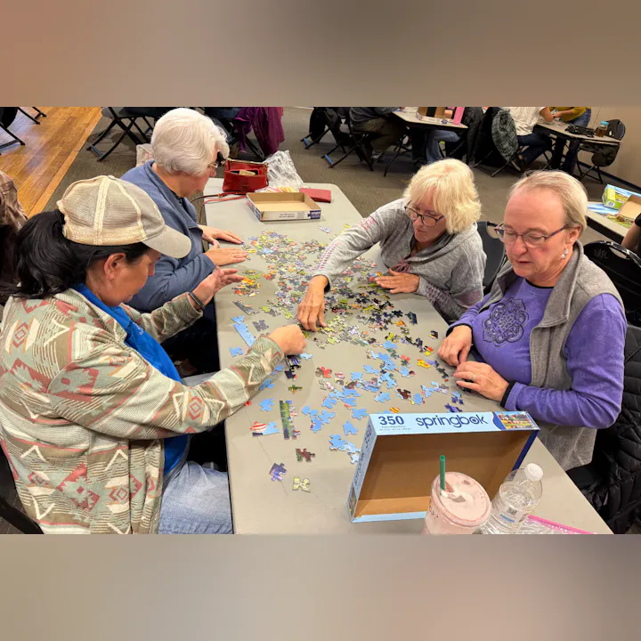 Four women are working on a jigsaw puzzle together at a table, surrounded by scattered puzzle pieces and drinks.