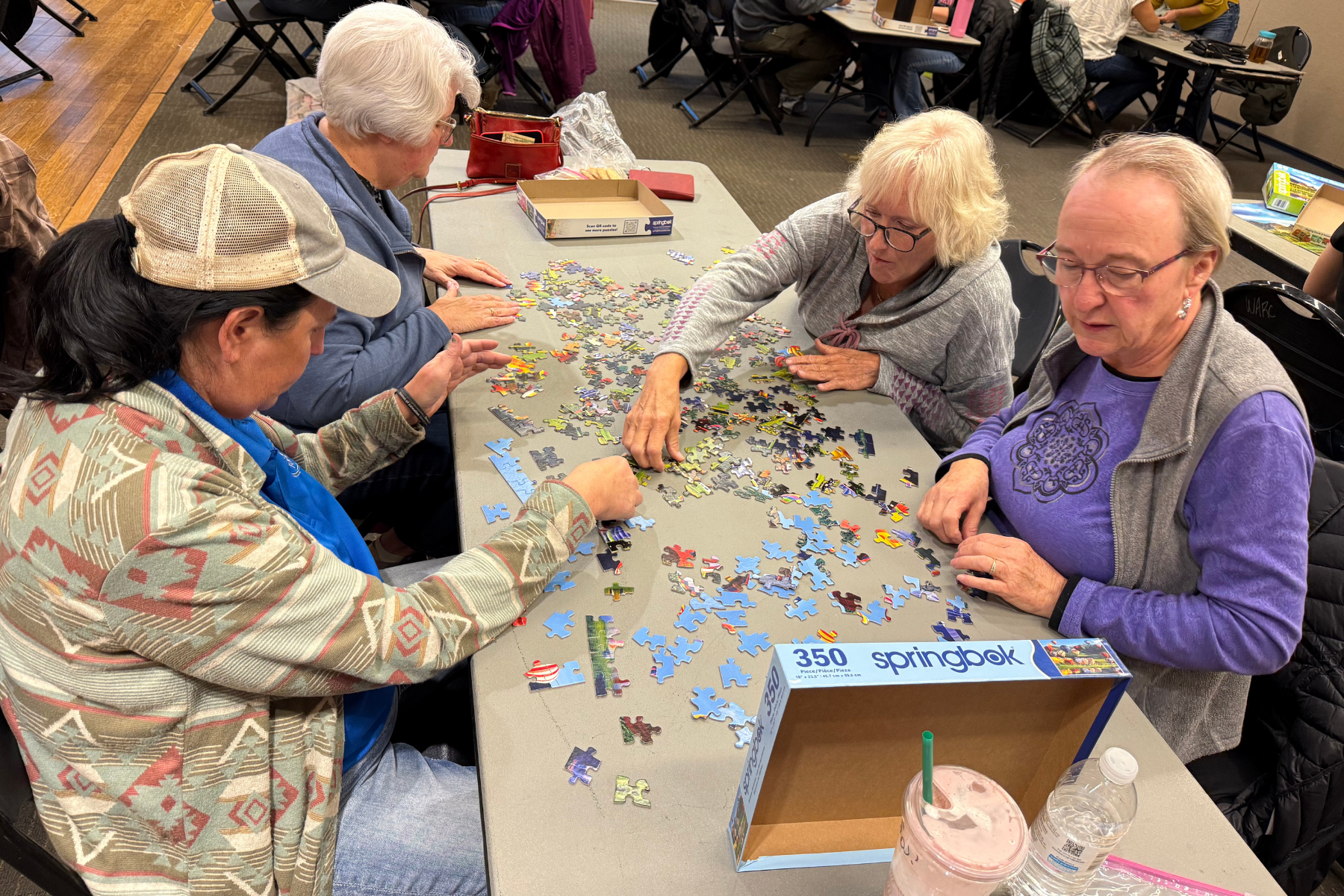 Four women are working on a jigsaw puzzle together at a table, surrounded by scattered puzzle pieces and drinks.