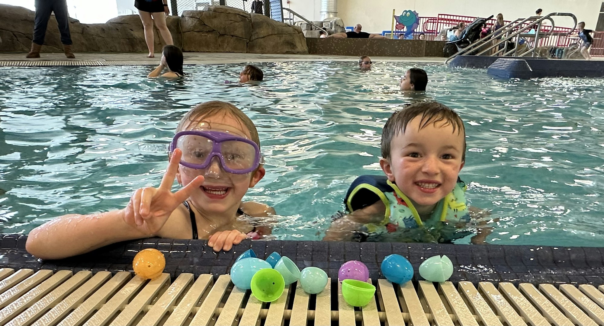 Two kids in a pool with colorful plastic eggs on the edge, one wearing goggles, both smiling.