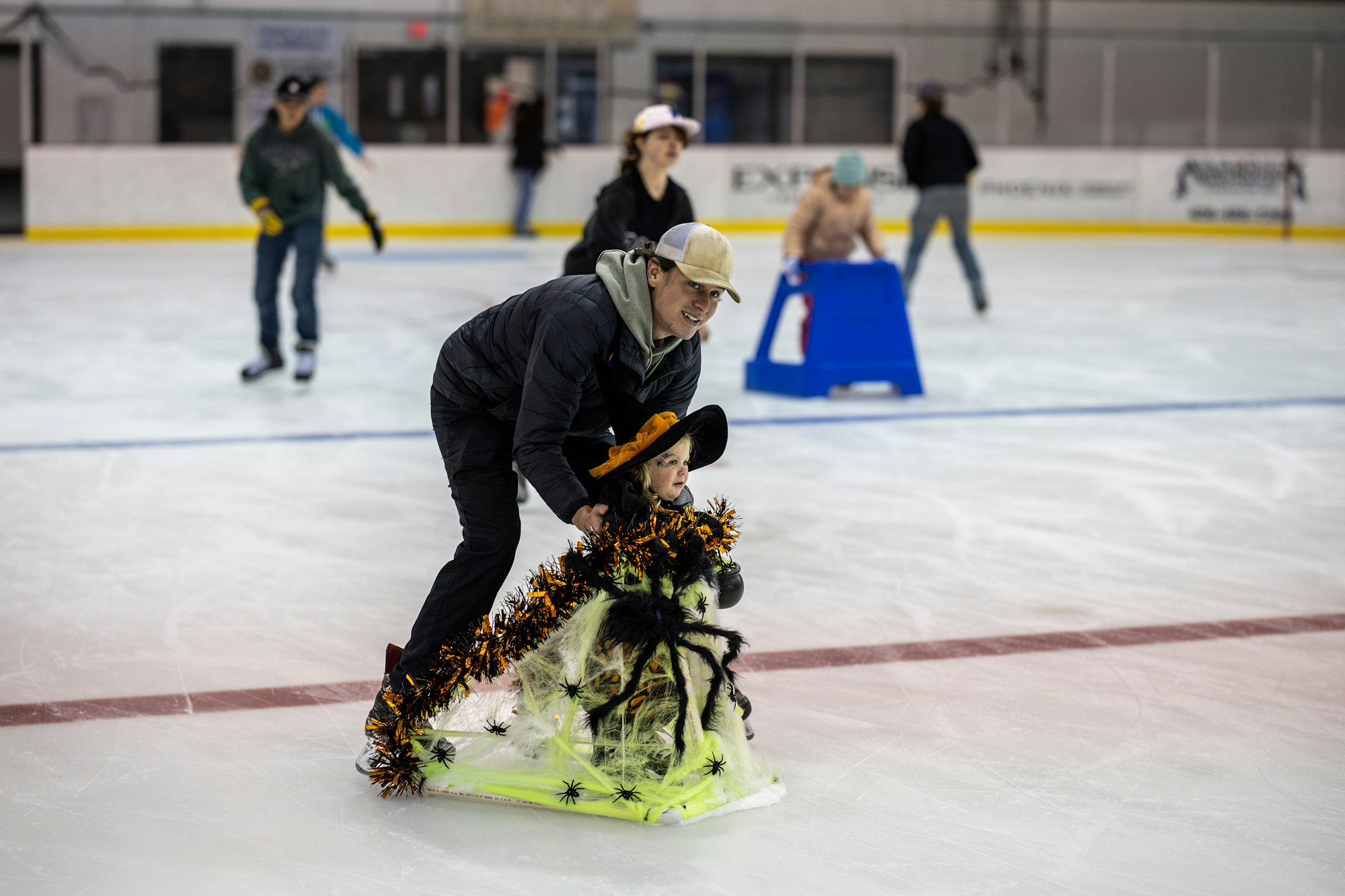 A person helps a child skate on ice, dressed in a festive Halloween outfit, while others skate in the background.