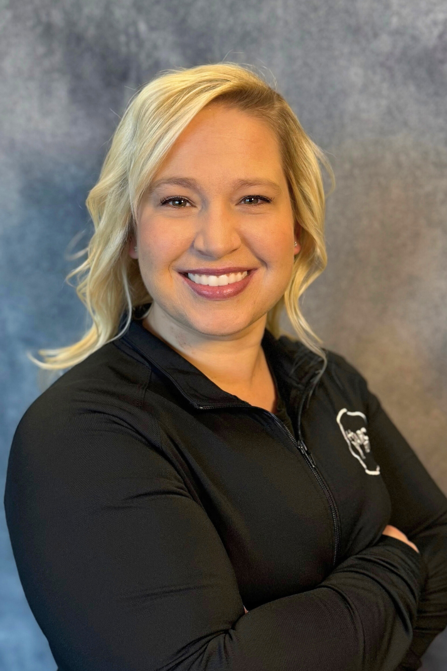 A person with blonde hair smiling, wearing a black jacket against a grey background.