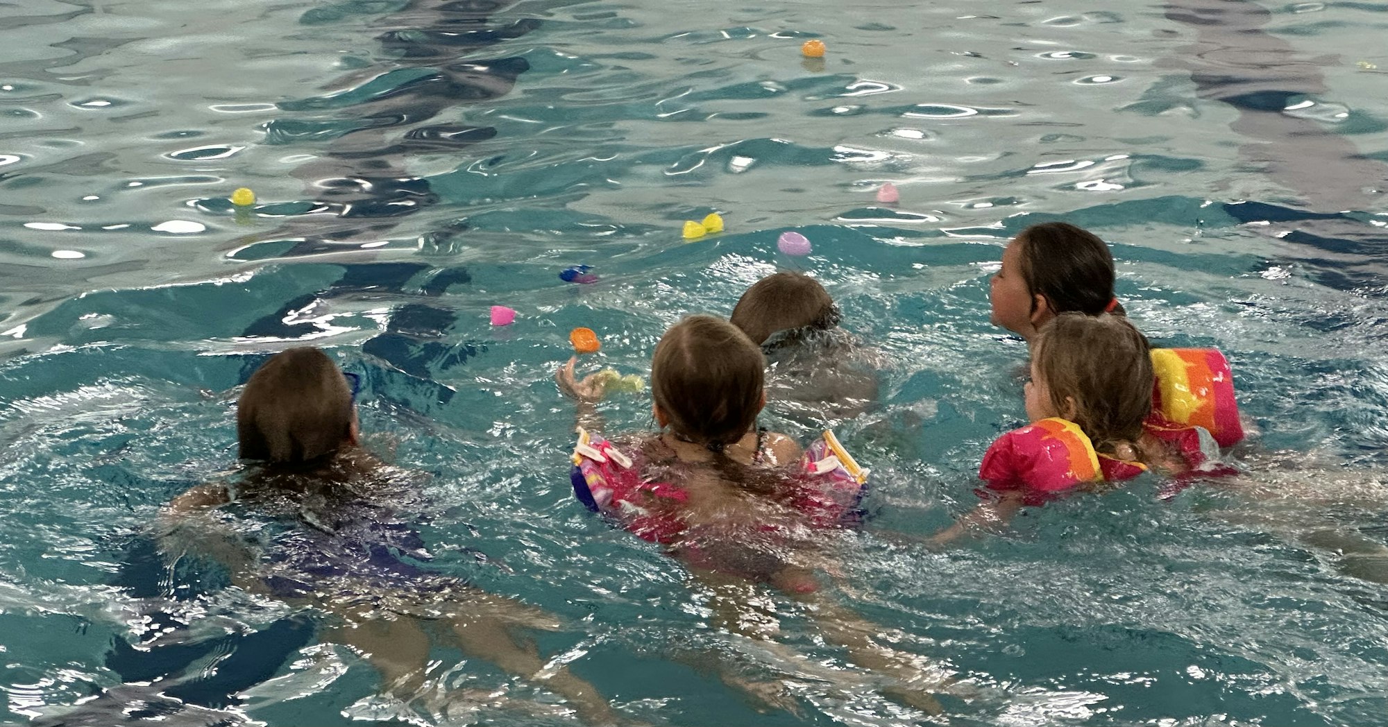 Children with floaties playing in a pool, with colorful toys floating around them.