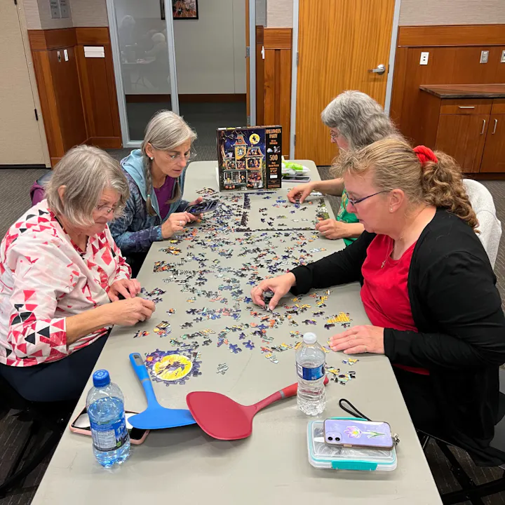 A group of four women is working together on a colorful jigsaw puzzle at a table, surrounded by snacks and water bottles.